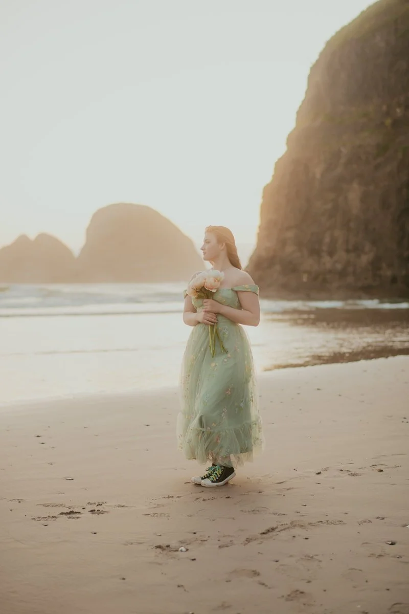 teen girl walking along the ocean holding peonies, wearing a green thrifted dress, and converse shoes