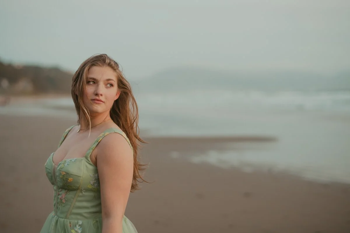 high school senior teen girl standing along the beach in oregon the wind blowing her hair, wearing a green dress.