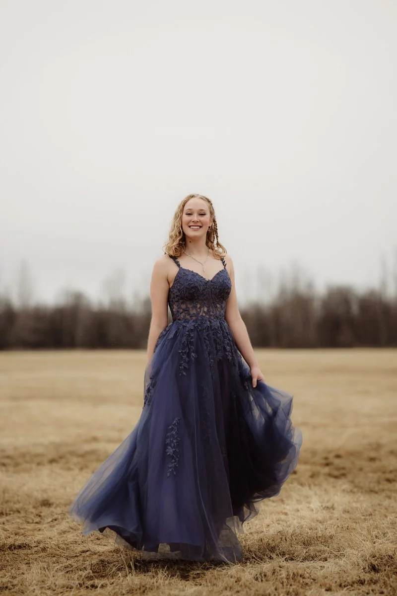 Girl standing in a field in Alaska for her prom senior portrait. Wearing a navy blue gown
