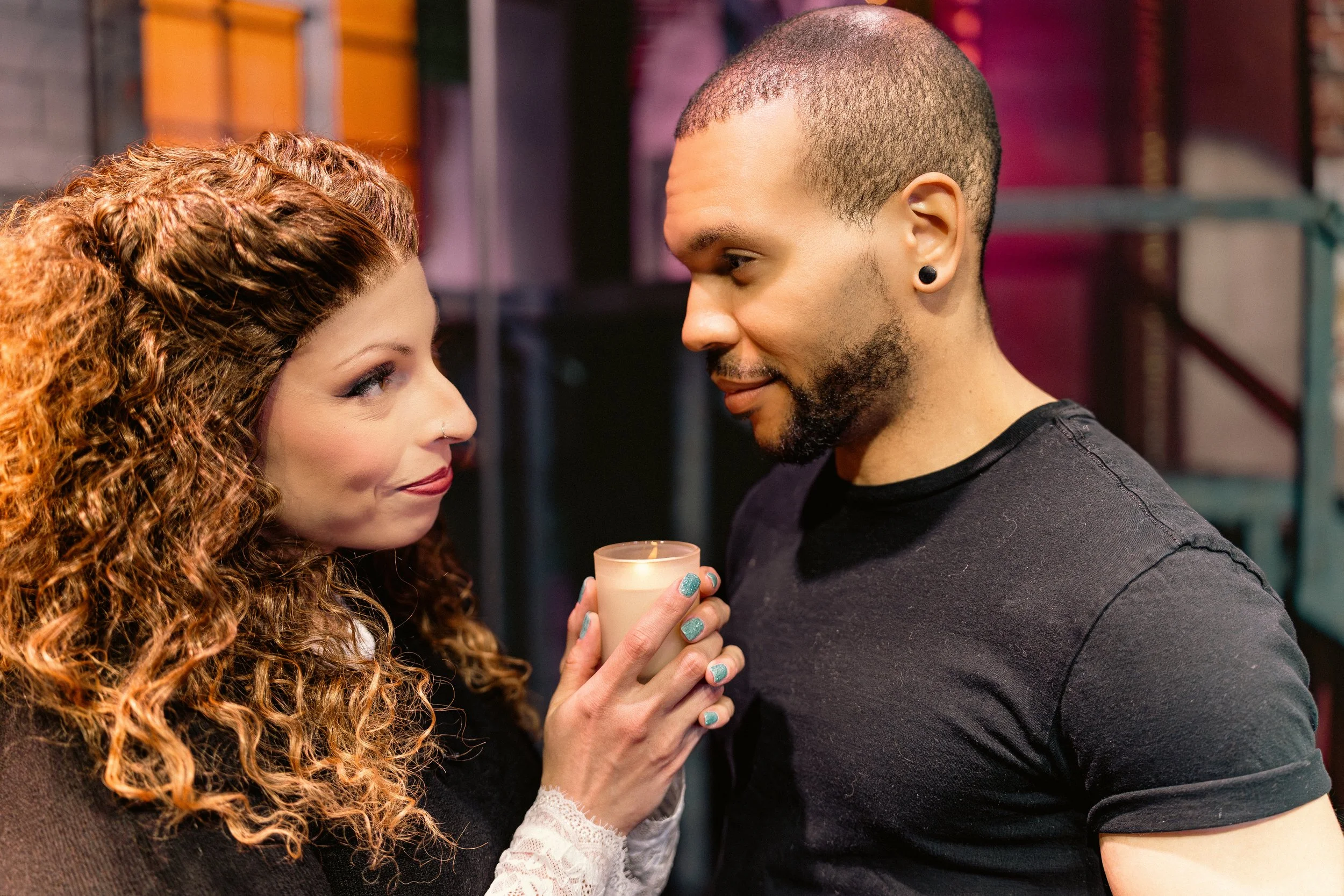 A woman with long, curly red hair holding a candle close to a man with a shaved head and beard, both smiling and looking at each other.