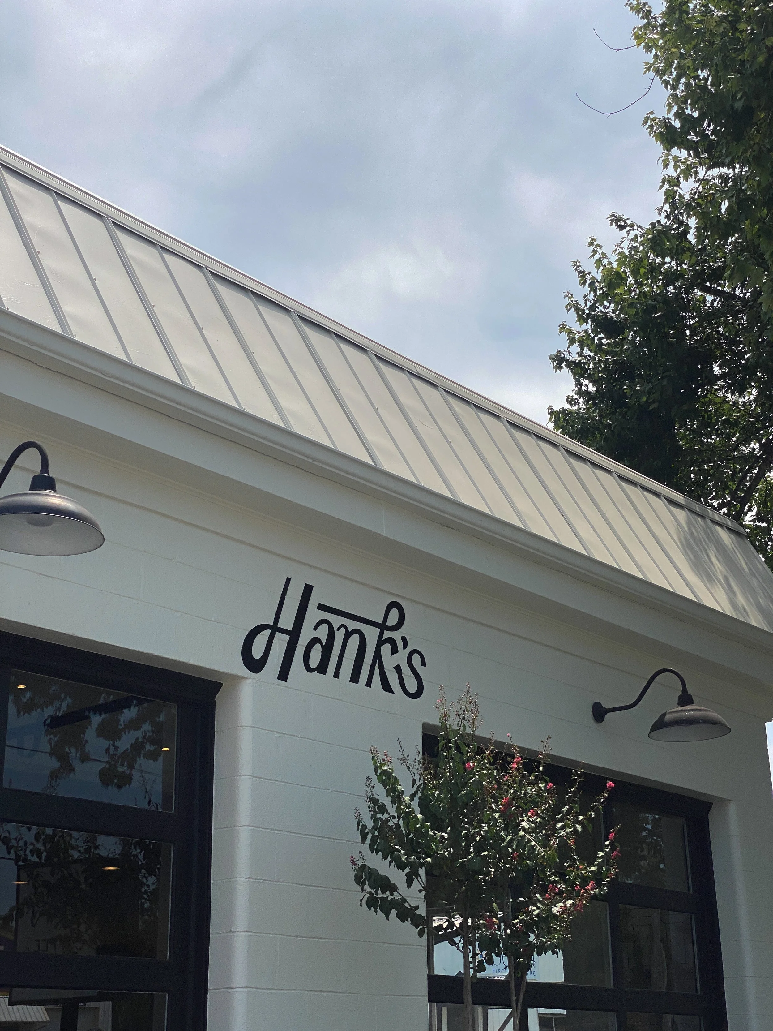 The exterior of a white building with the sign 'Hank's' on the wall, two black wall-mounted lamps, a tree in front, and a cloudy sky above.