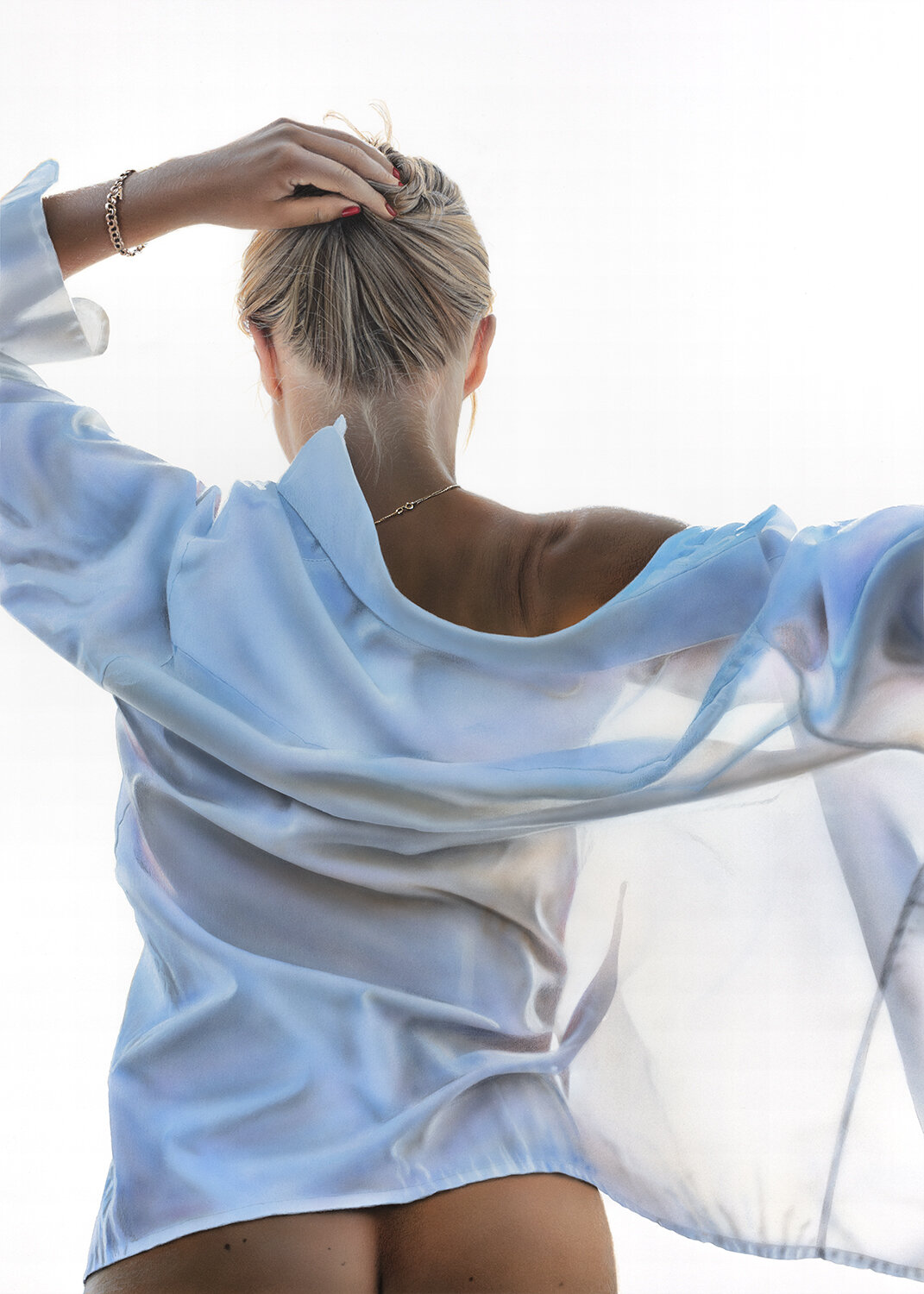 Hyperrealistic painting by Johannes Wessmark. Back view of a woman with blonde hair in a messy bun, wearing a shiny, light blue satin blouse. She is touching her head with one hand, and the photo has a bright, white background.