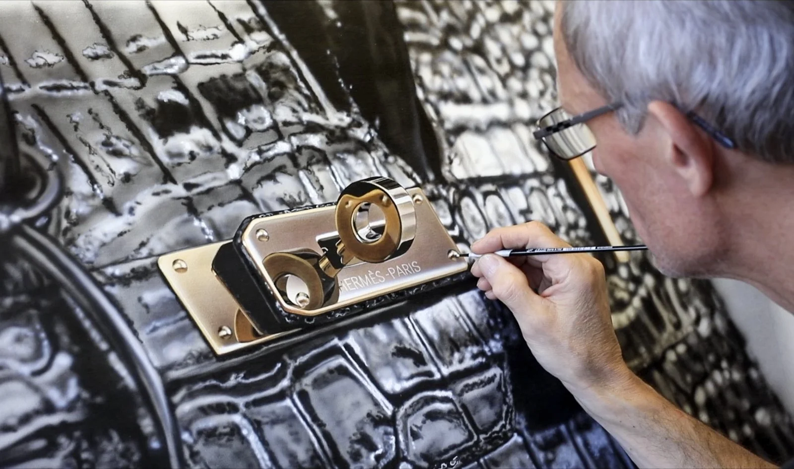 An artisan painting or restoring a black crocodile leather handbag with a gold and silver Hermès Paris logo and lock hardware, using a fine brush for detailing.