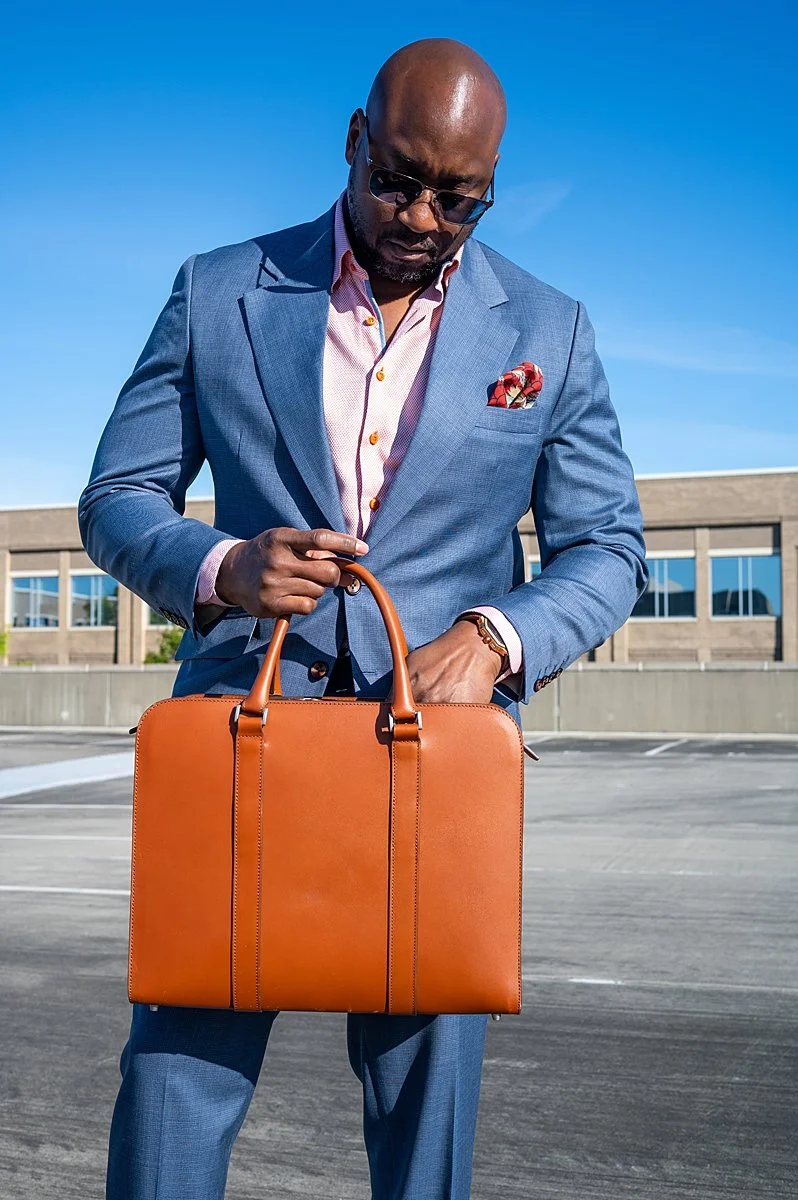 A man in a blue suit and sunglasses holding a brown leather briefcase in an outdoor parking lot on a clear day.