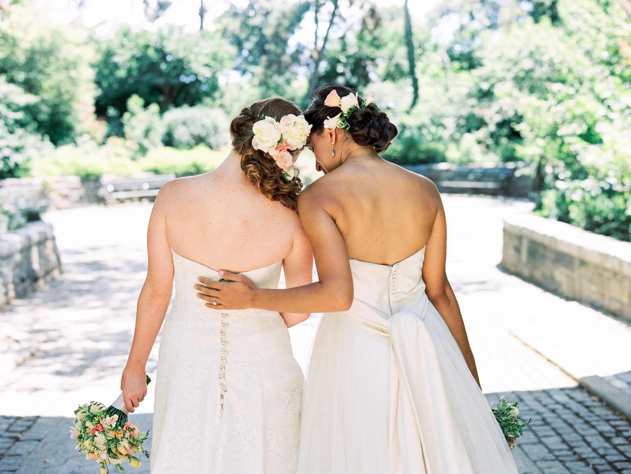 LGBTQ+ newlywed couple in wedding dresses holding bouquets, walking down cobblestone street i NYC.
