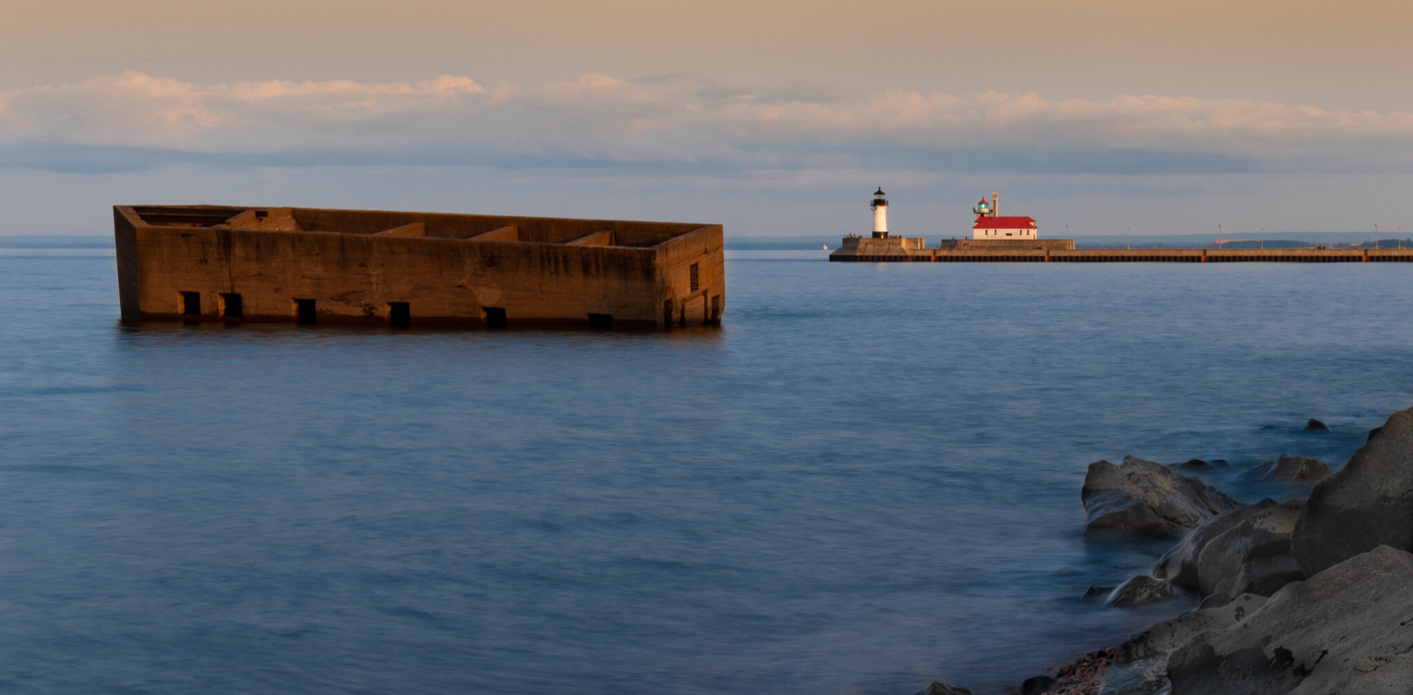 A lighthouse and a small building on a pier in the distance, with a large submerged wooden structure in the water in the foreground, rocks along the shore, calm water, and a cloudy sky.