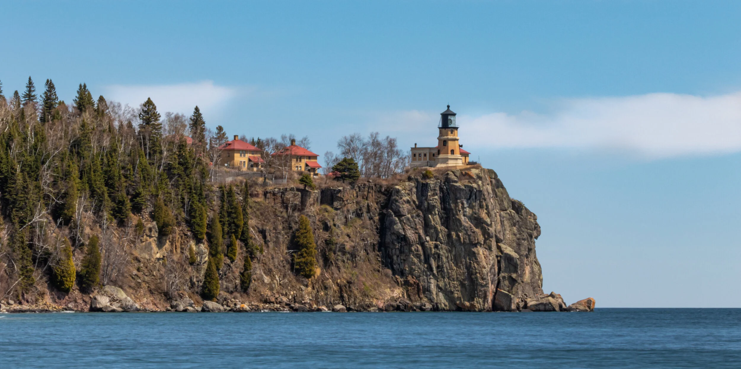 A lighthouse on a cliff overlooking the ocean with houses and trees nearby, under a blue sky with clouds.