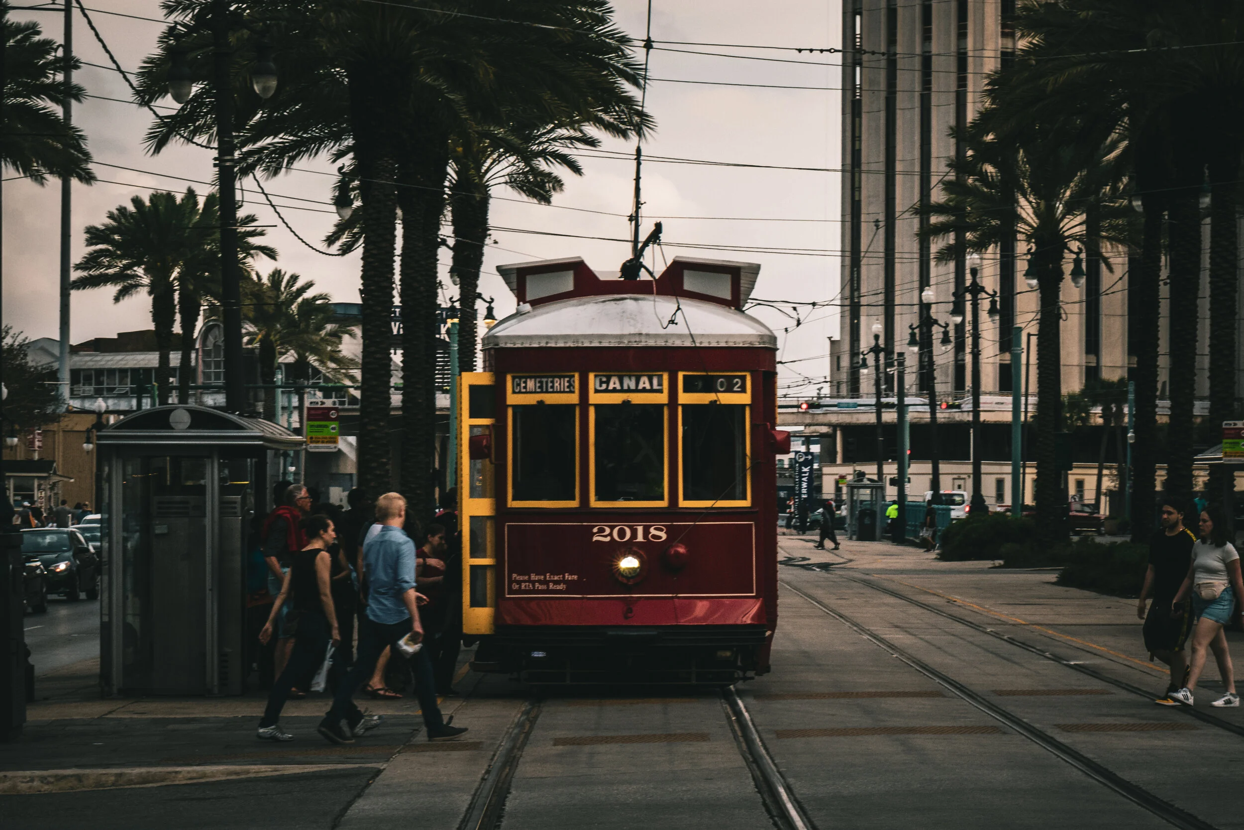 A red trolley with the sign 'CEMETERIES CANAL 02' is stopped at a street crossing with pedestrians walking across the tracks. Palm trees and city buildings are visible in the background amid overcast skies.