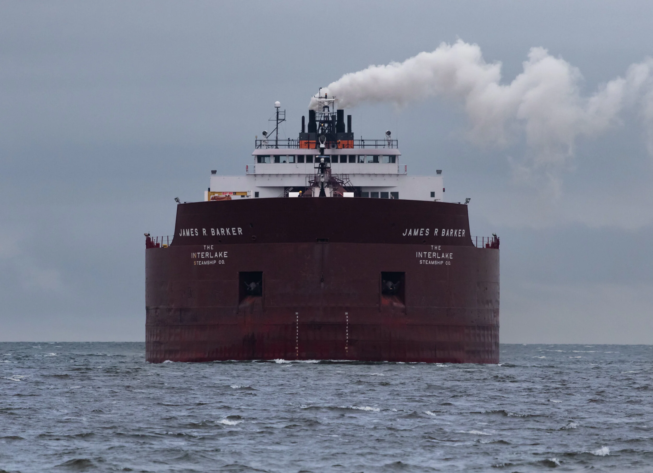 A large cargo ship named James R Barker on the water, with gray overcast sky and smoke coming from the smokestack.