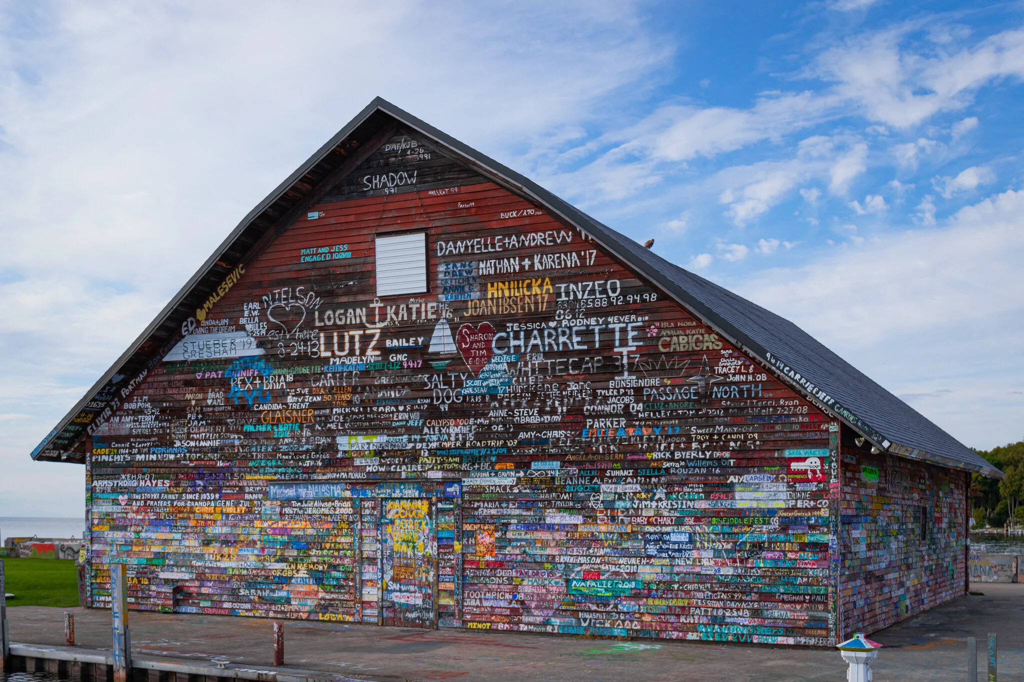 A large wooden building covered with colorful painted names and messages, situated outdoors with a partly cloudy sky in the background.