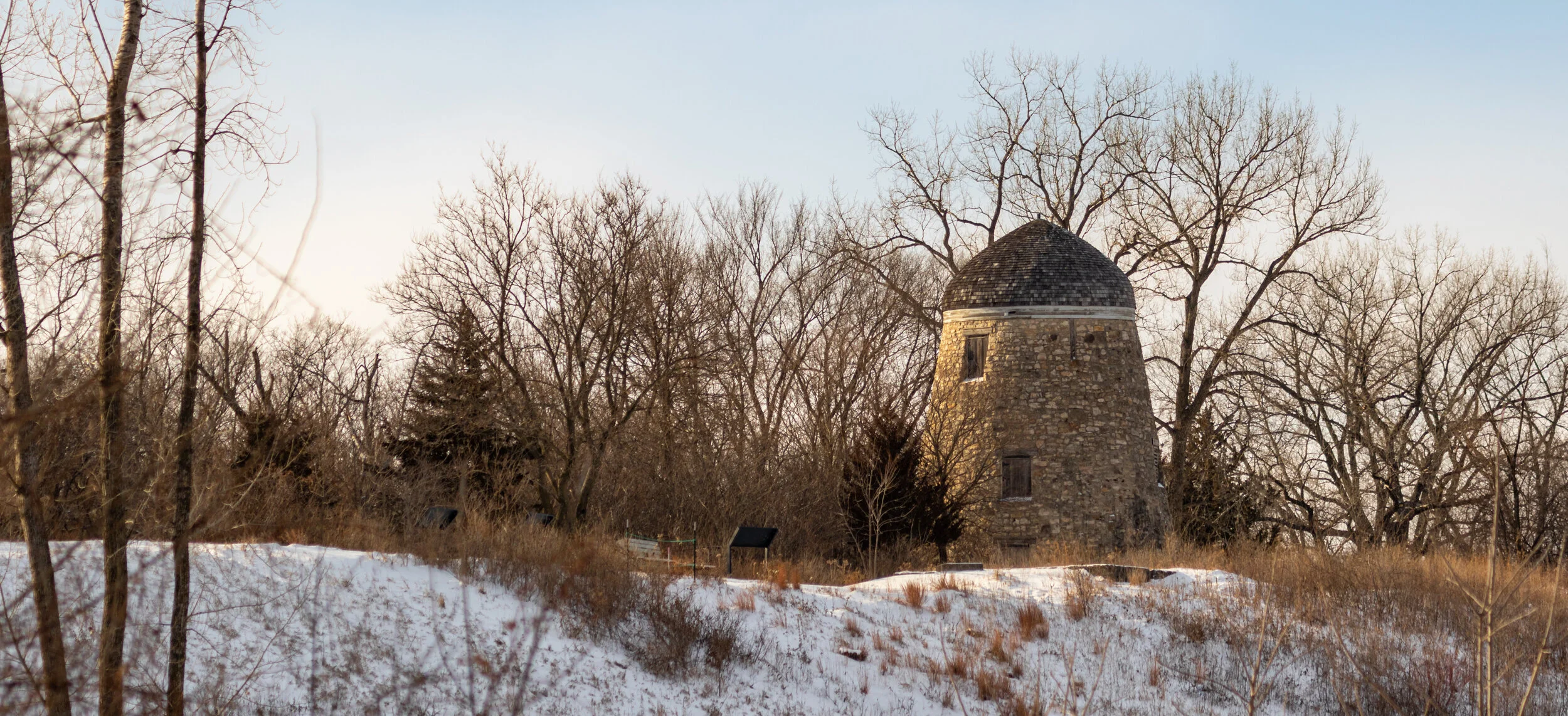 A stone round tower with a domed roof in a Winter landscape with leafless trees and snow on the ground.
