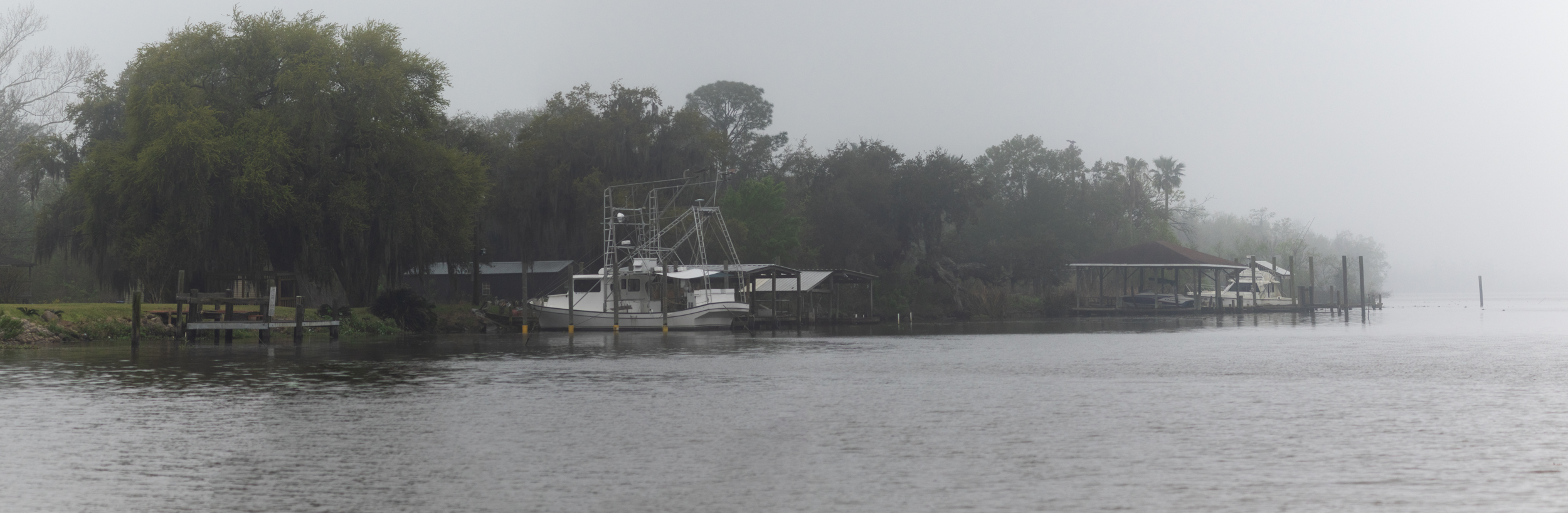 A foggy waterfront scene with boats docked along a shoreline with trees and small structures.