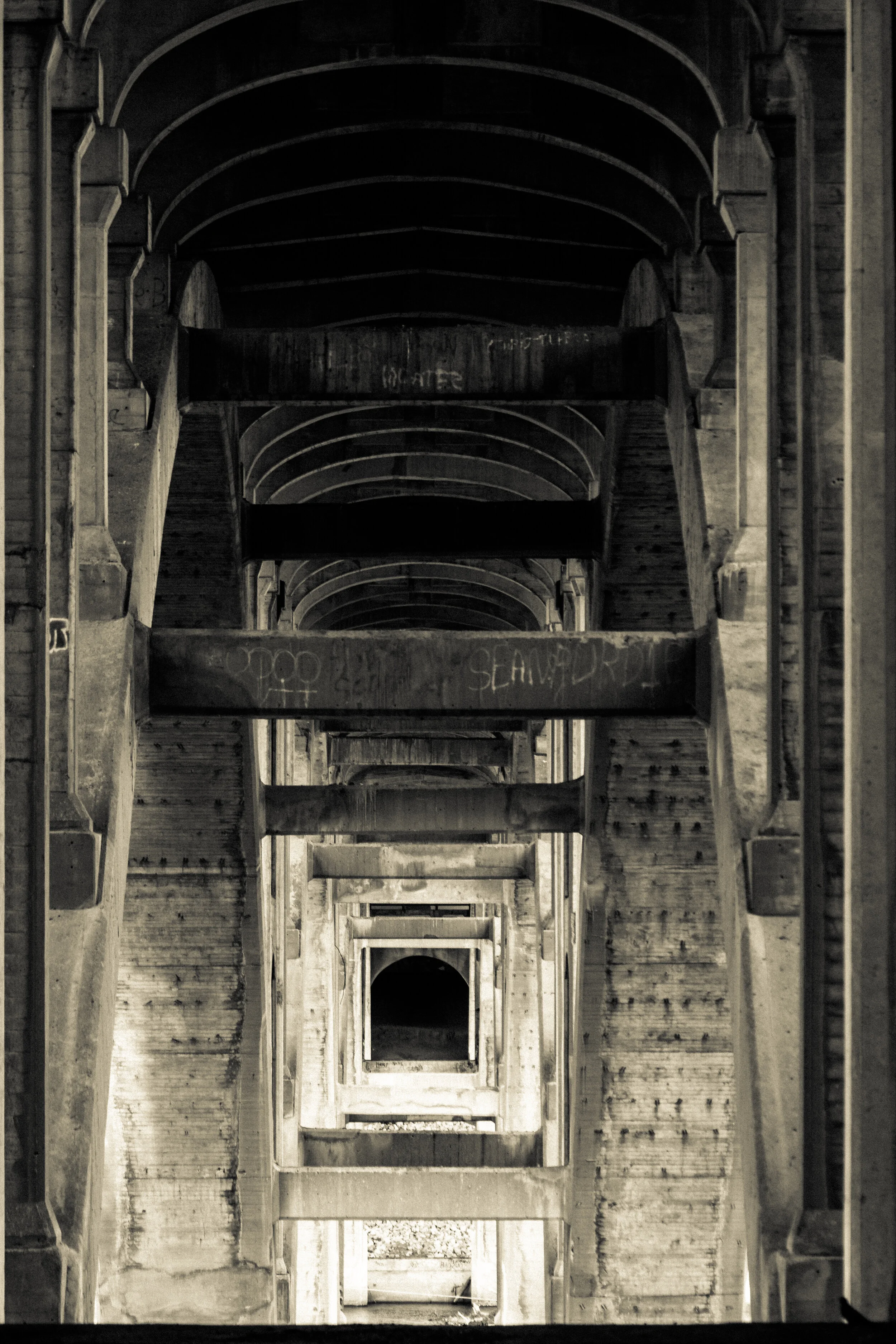 A black-and-white photo of multiple rectangular, wooden beams and arches creating a tunnel-like structure, with some graffiti visible on the beams.