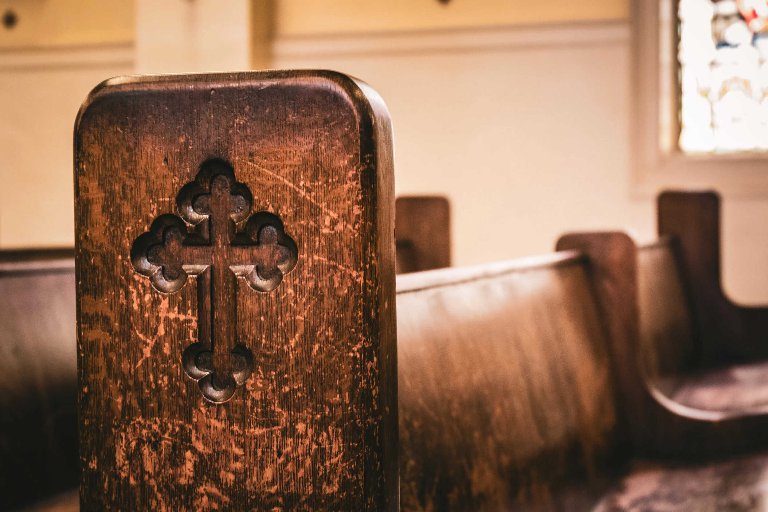 Close-up of a wooden church pew with a carved cross on its end, in a softly lit church interior with stained glass window in the background.