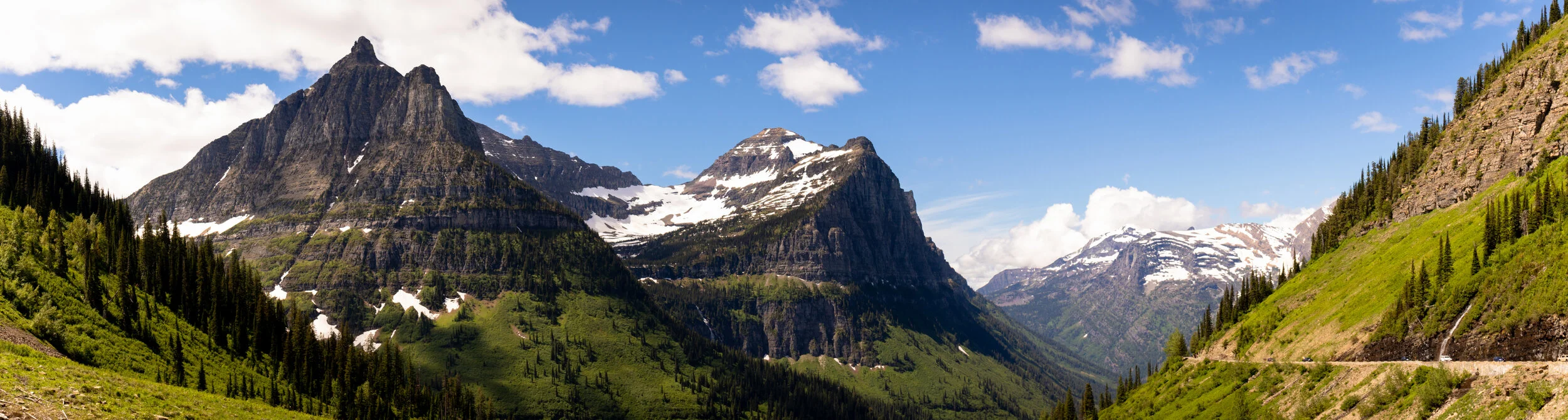 Mountain range with snow-capped peaks, green valleys, and forested slopes under blue sky with scattered clouds.