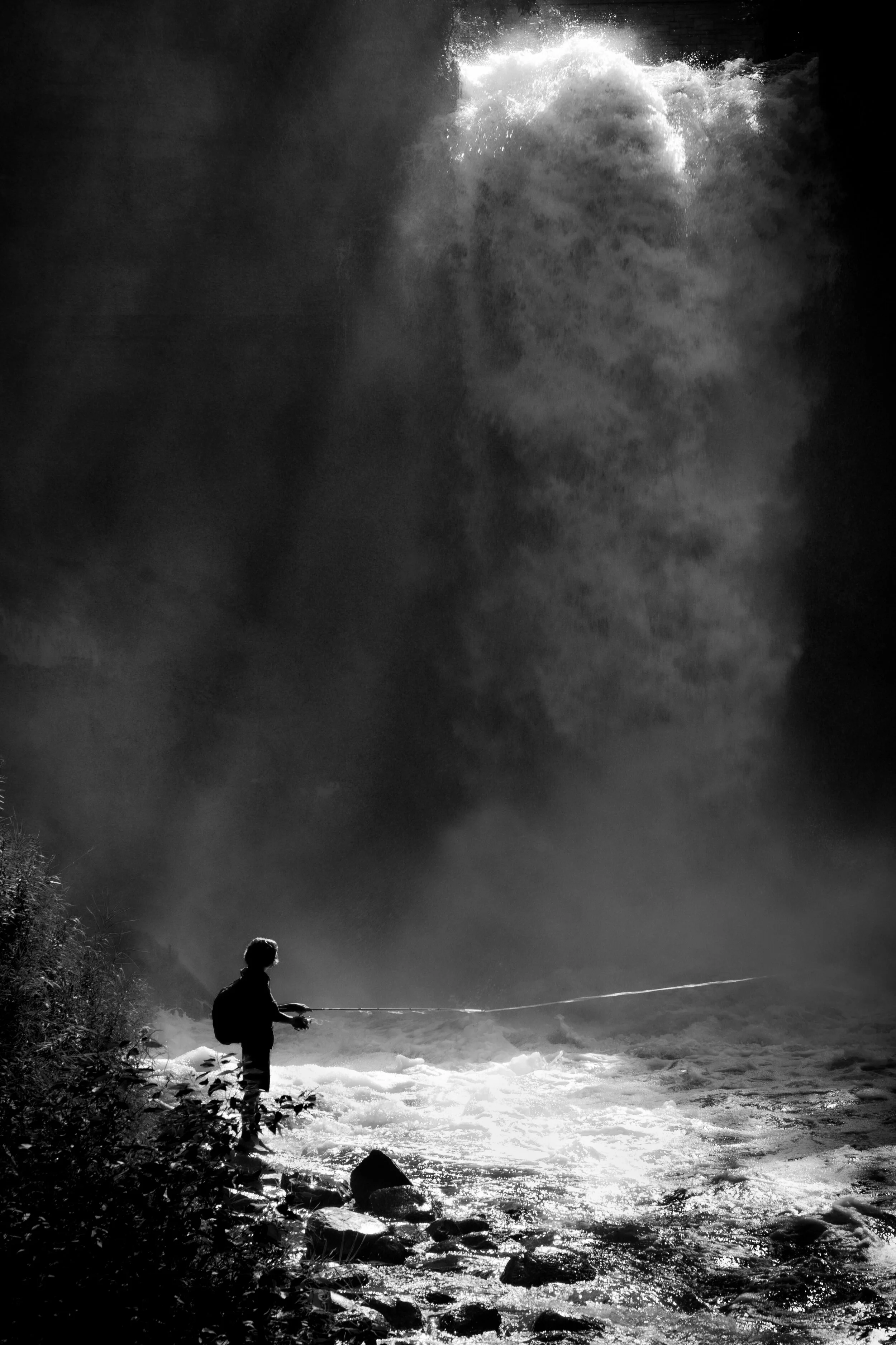 Silhouette of a person fishing by a river with waterfall in the background, black and white