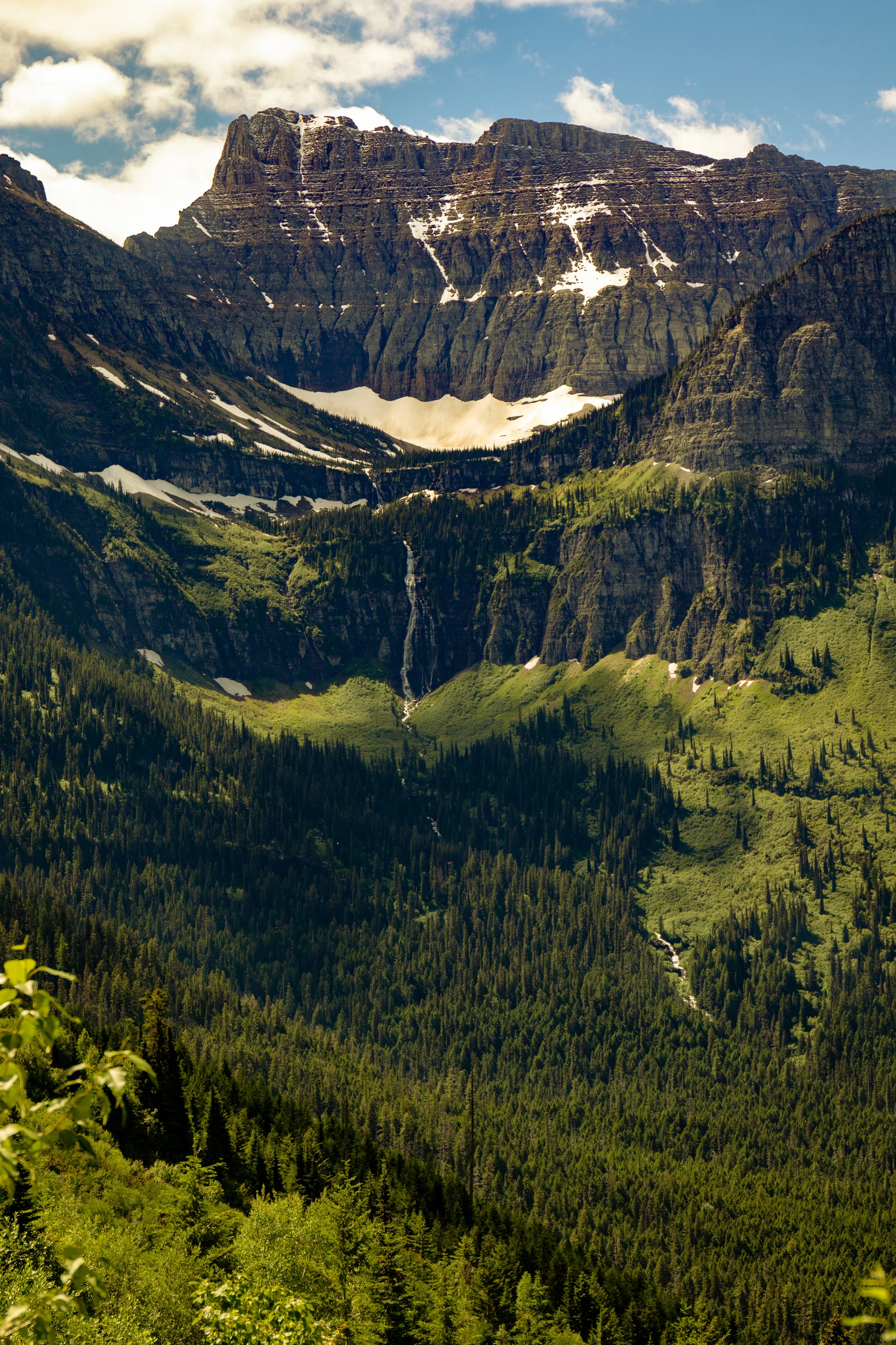 A tall mountain with snow on its peak and slopes, surrounded by lush green trees in a forested valley with a small waterfall flowing down the hillside.