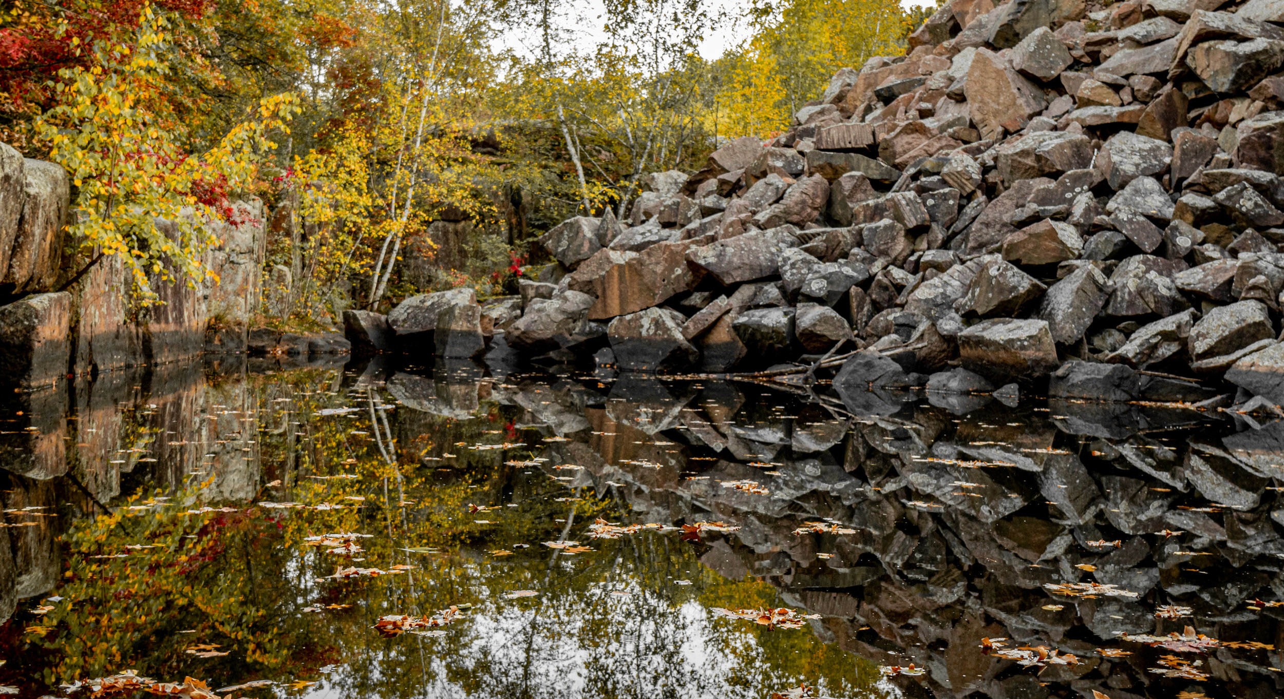 A rocky hillside beside a calm body of water reflecting the rocks and colorful autumn trees.
