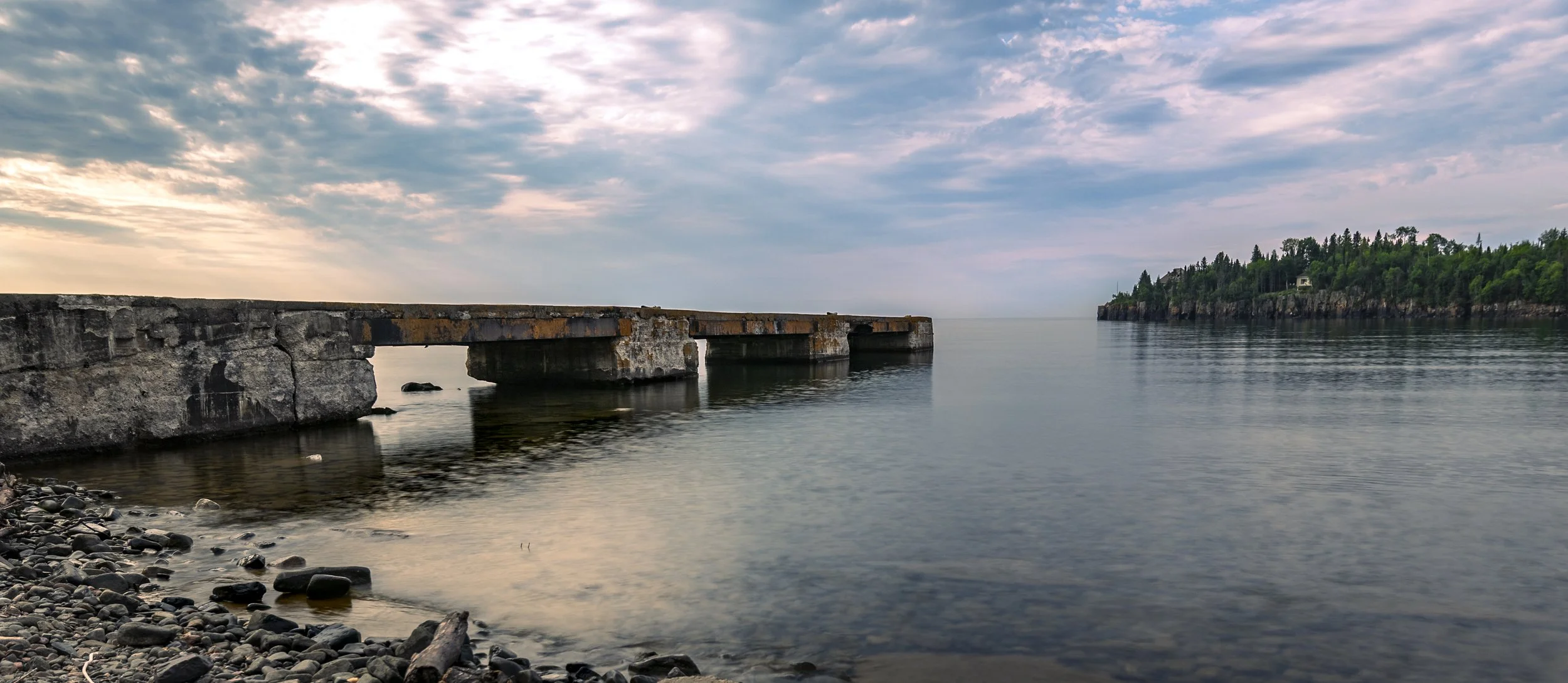 Calm body of water with an old, rusted stone pier extending into the water, a tree-covered island in the distance, and a partly cloudy sky at sunset.