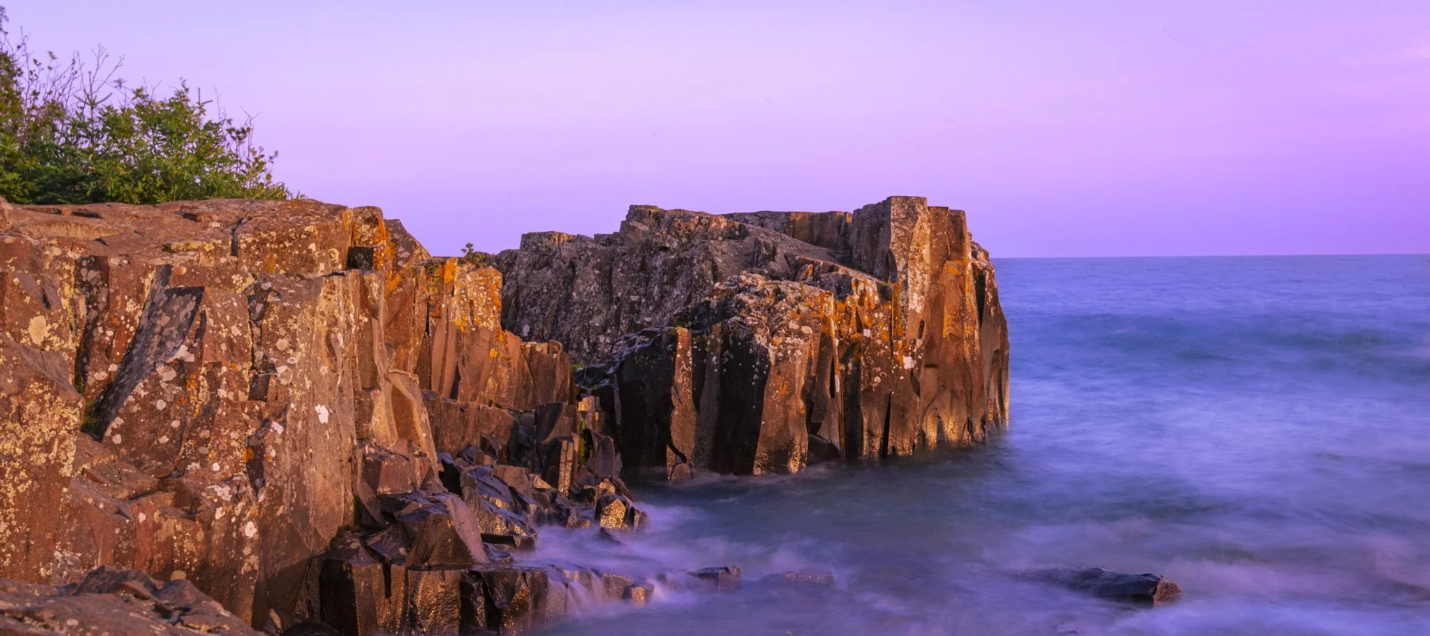 Rocky coastline at sunset with purple sky and calm ocean water.