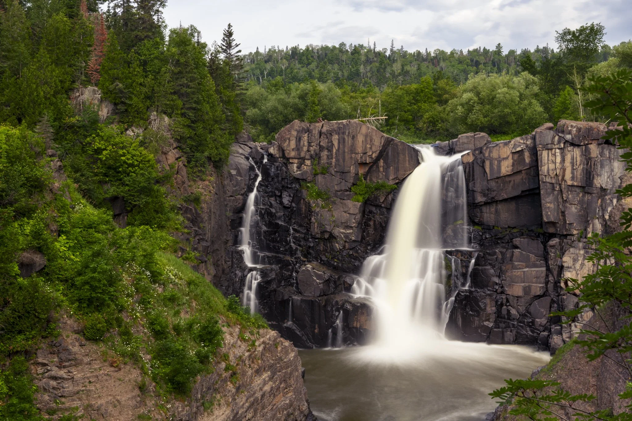 A waterfall cascading over dark rocks surrounded by lush green trees and plants in a forested landscape.