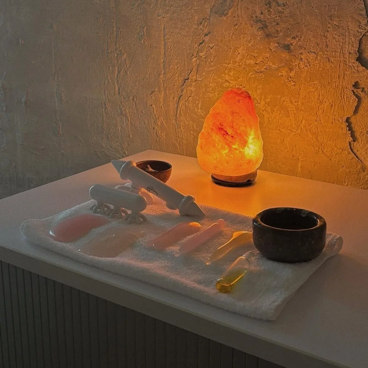 A salt lamp on a table casting warm light. A self-care or massage setup with massage stones, a massage roller, and small bowls, all resting on a white towel.