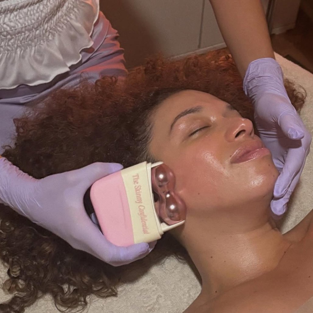 A woman receiving a facial treatment with a device on her jawline, lying on a treatment bed with closed eyes, while a professional wearing purple gloves conducts the procedure.