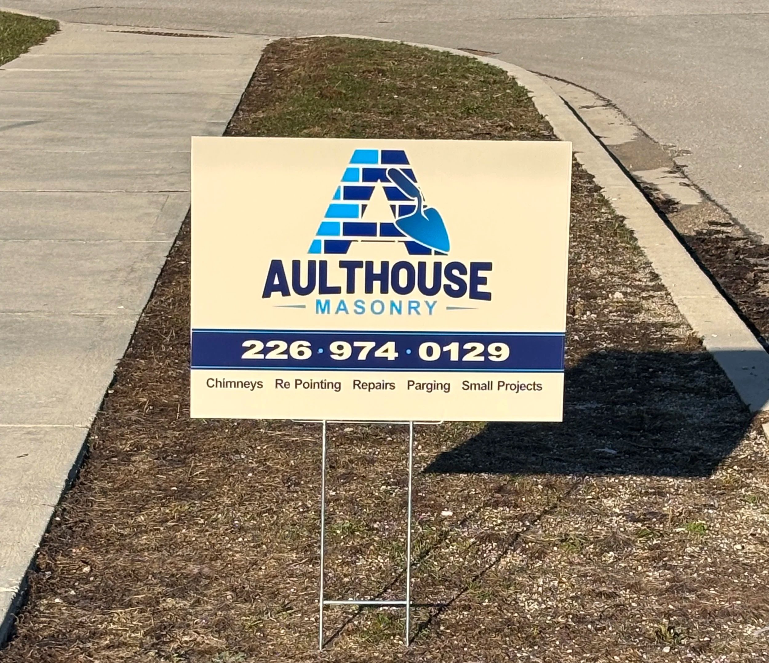 A sign in front of a sidewalk and some grass. The sign has a logo with bricks and a trowel and says: 'AULT HOUSE MASONRY 226 974 0129 Chimneys Re Pointing Repairs Parging Small Projects' - Signupsigns - Neustadt.