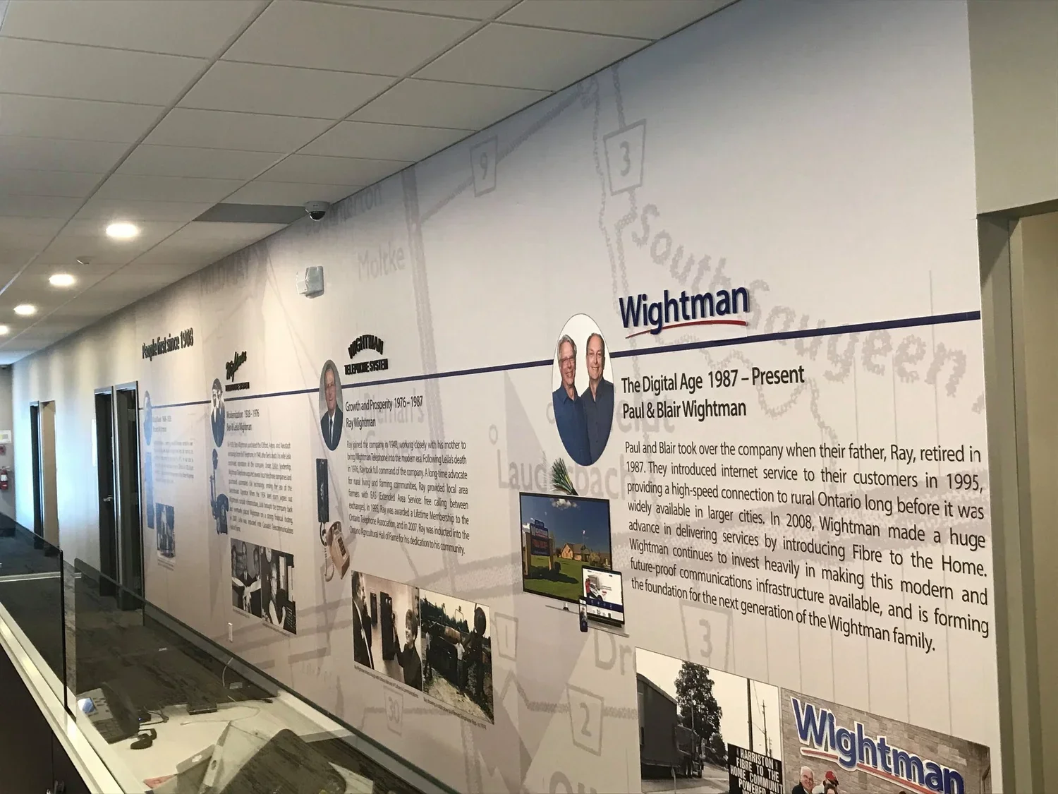A wall display featuring the history of Wightman, a telecommunications company, with text and photos of key figures including Paul and Blair Wightman, along with images of company milestones and local community activities.