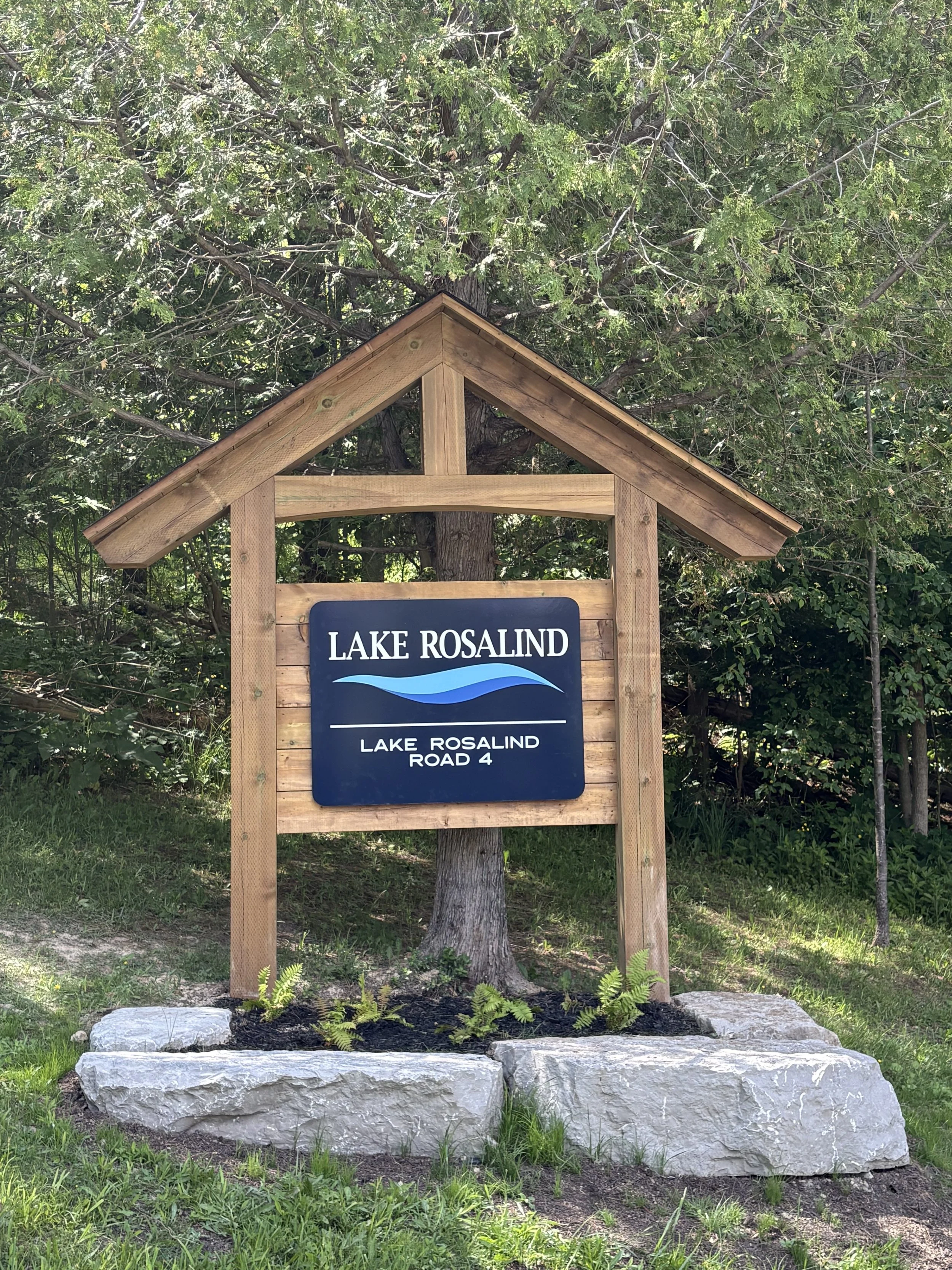 Wooden sign with a roof that reads "Lake Rosalind, Lake Rosalind Road 4", situated on a rock base, against a background of trees and greenery.