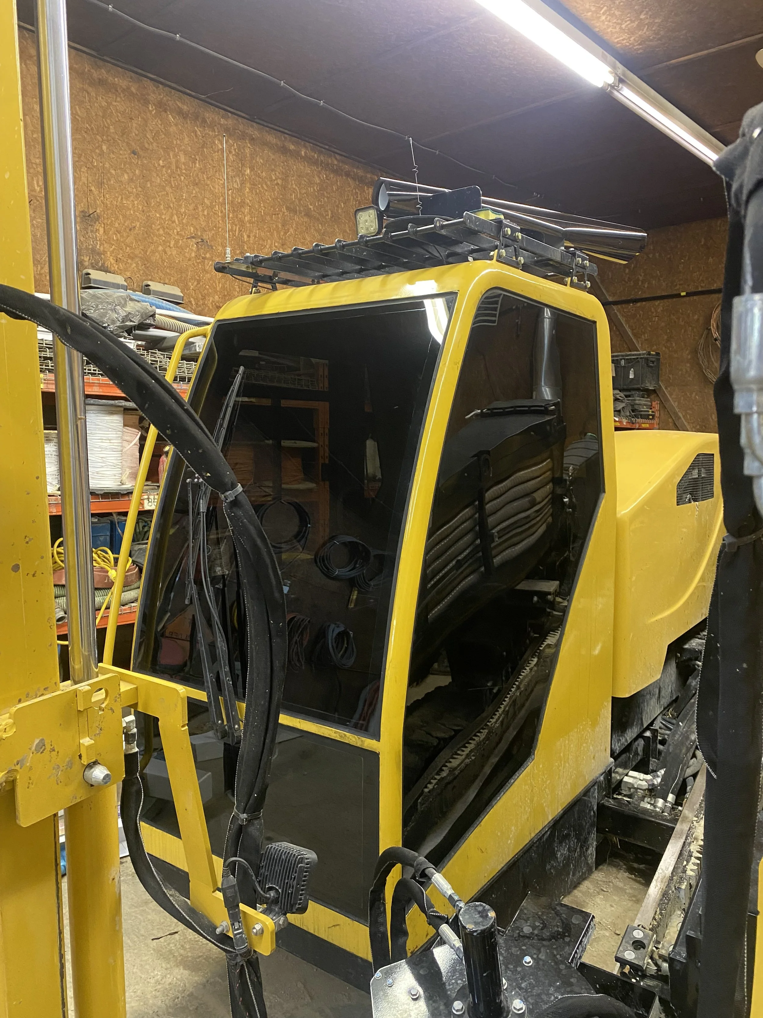 The image shows a yellow construction vehicle, likely a small excavator or skid steer, inside a workshop or garage. The vehicle's cab is visible, with a black interior and large glass windows. On top of the vehicle, there's a roof rack with various tools or equipment attached. The background includes shelves with storage bins and other tools, and the walls are made of wood paneling. The lighting is fluorescent.