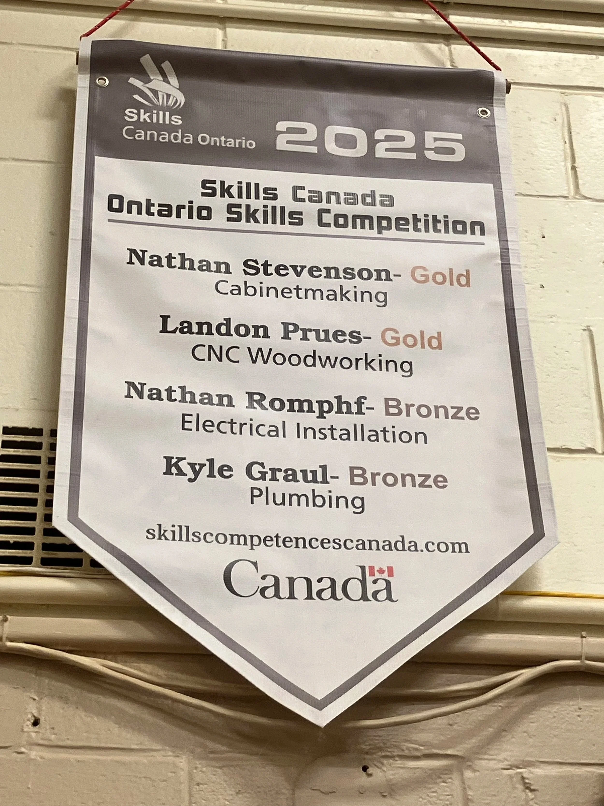 A printed banner hanging on a wall, featuring Skills Canada Ontario 2025 and listing competitors Nathan Stevenson for Cabinetmaking, Landon Prues for CNC Woodworking, Nathan Romphf for Electrical Installation, and Kyle Graul for Plumbing, Signupsigns