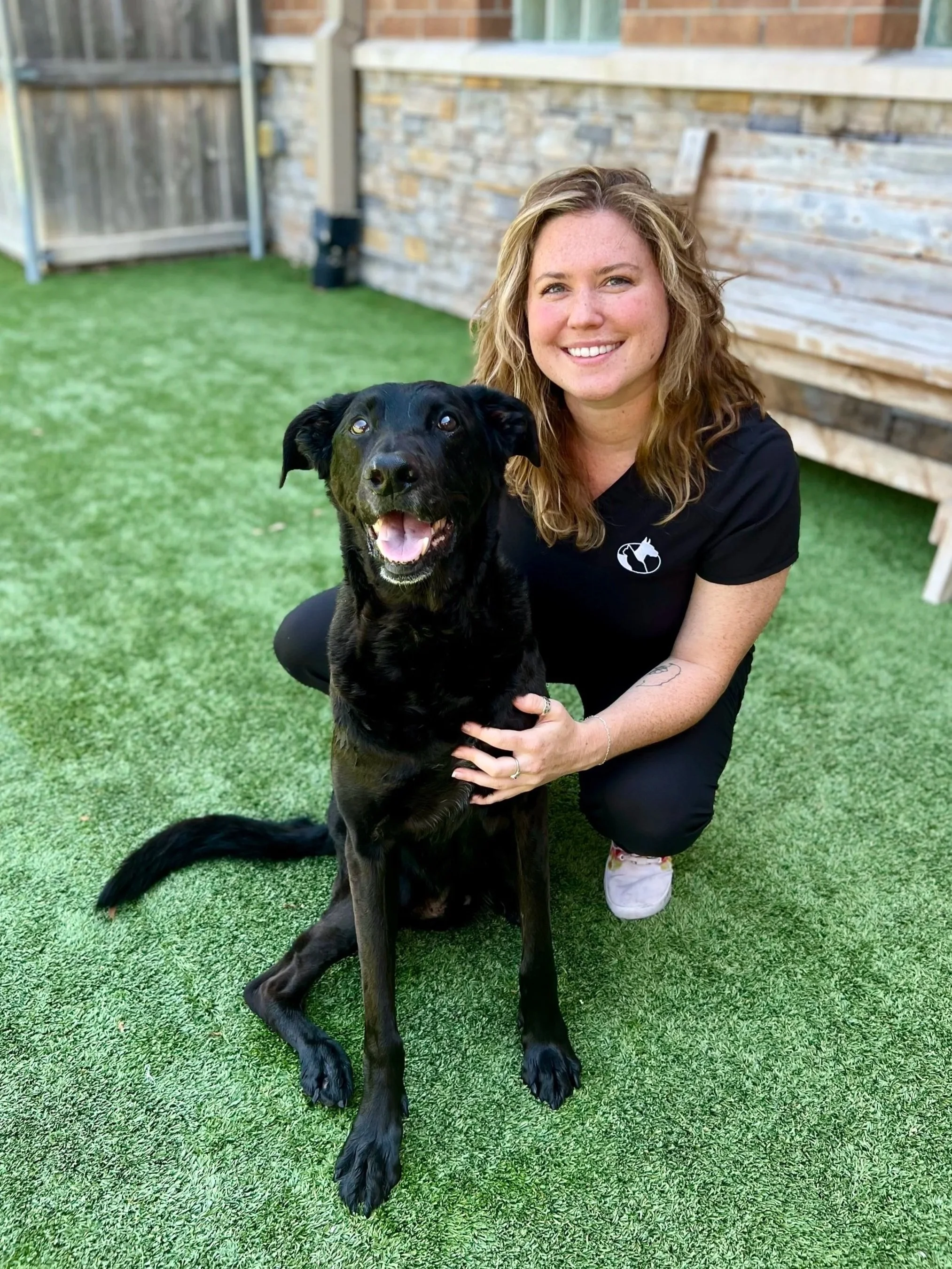A woman kneeling on grass next to a black dog, smiling at the camera.