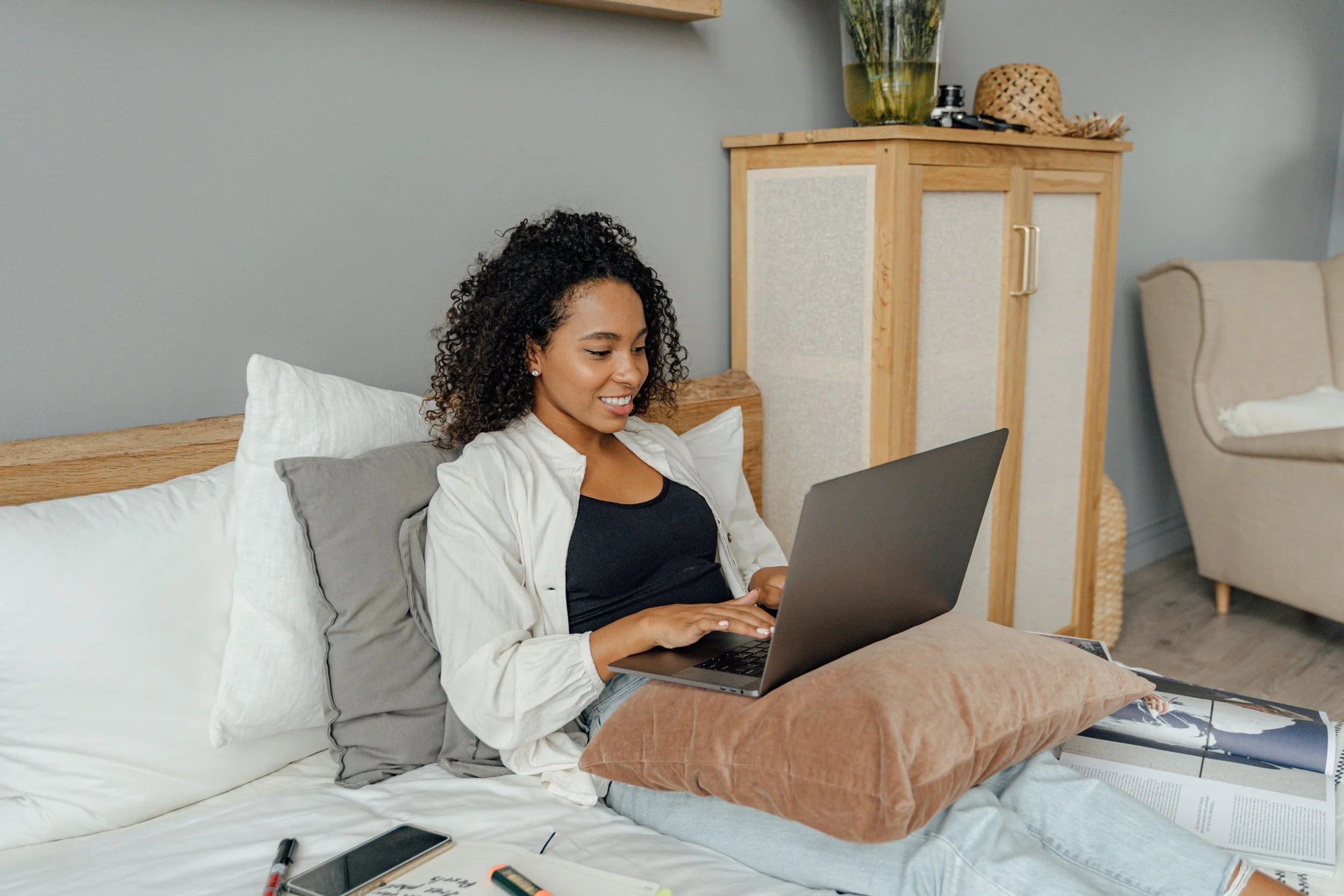 A woman with curly hair sitting on a bed, working on a laptop, surrounded by papers, notebooks, markers, and a smartphone.