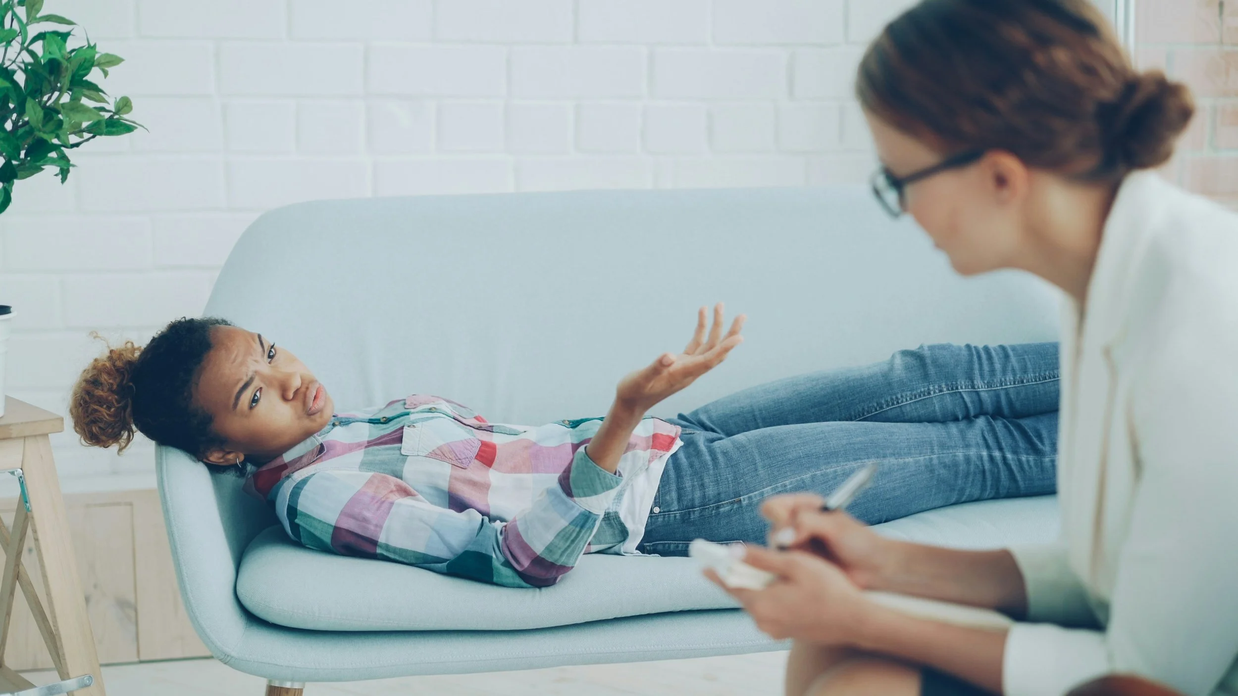 A woman lying on a light-colored couch, talking to a mental health professional in an office setting.