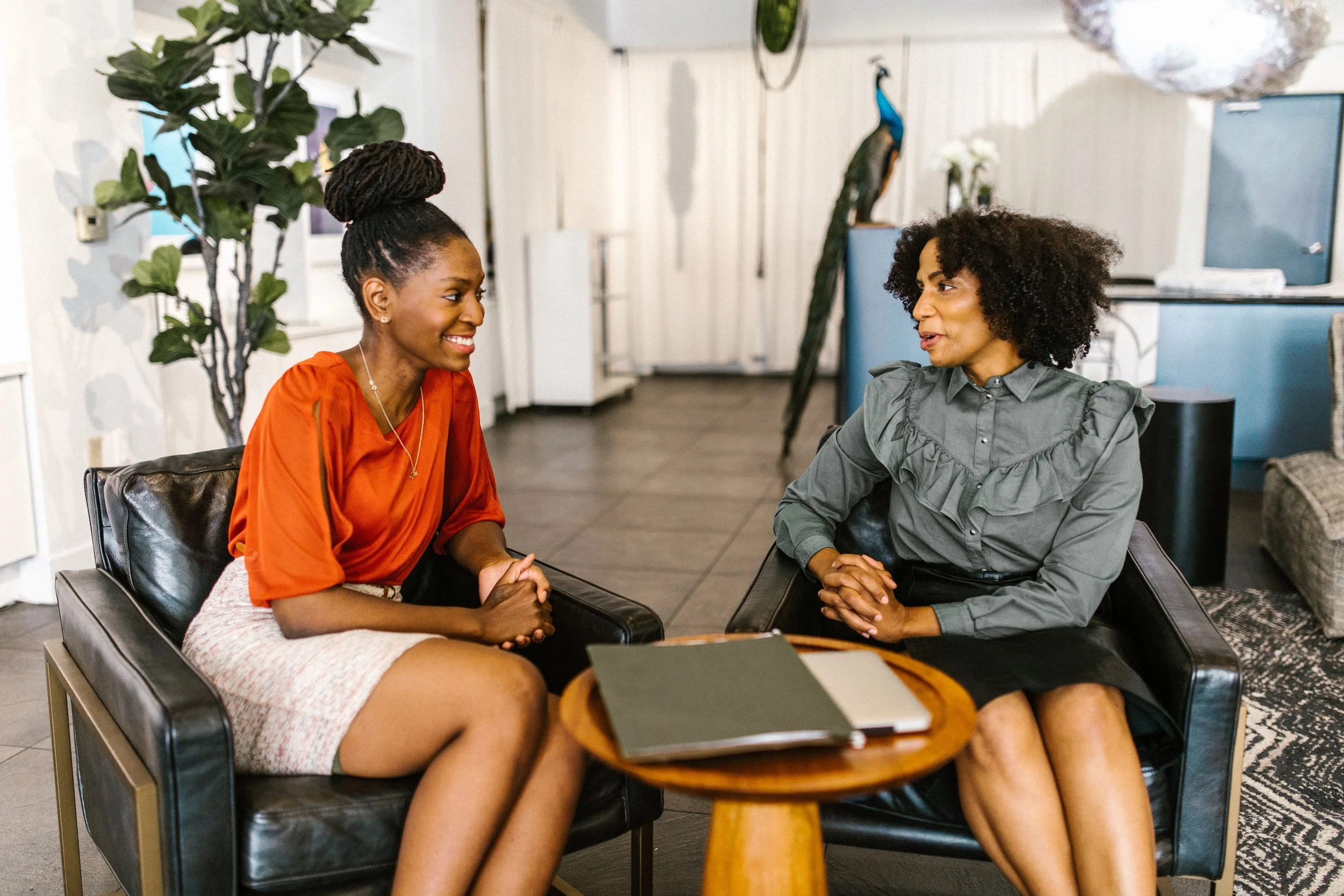 Two women are sitting at a table, having a conversation. One is wearing an orange top and a skirt, and the other is wearing a gray shirt. They are smiling and appear to be engaged in a friendly discussion. The setting is indoors with furniture and plants visible in the background.