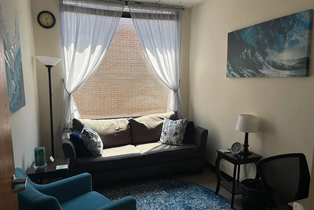 Living room with a large window, sheer curtains, a gray sofa with decorative pillows, a small side table with a lamp and clock, a blue armchair, a round black table, a floor lamp, and ocean-themed artwork on the walls.