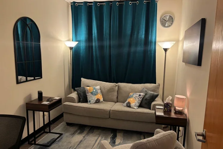 Living room with beige couch, dark wood side tables, blue curtains, wall clock, wall art, and lamps.