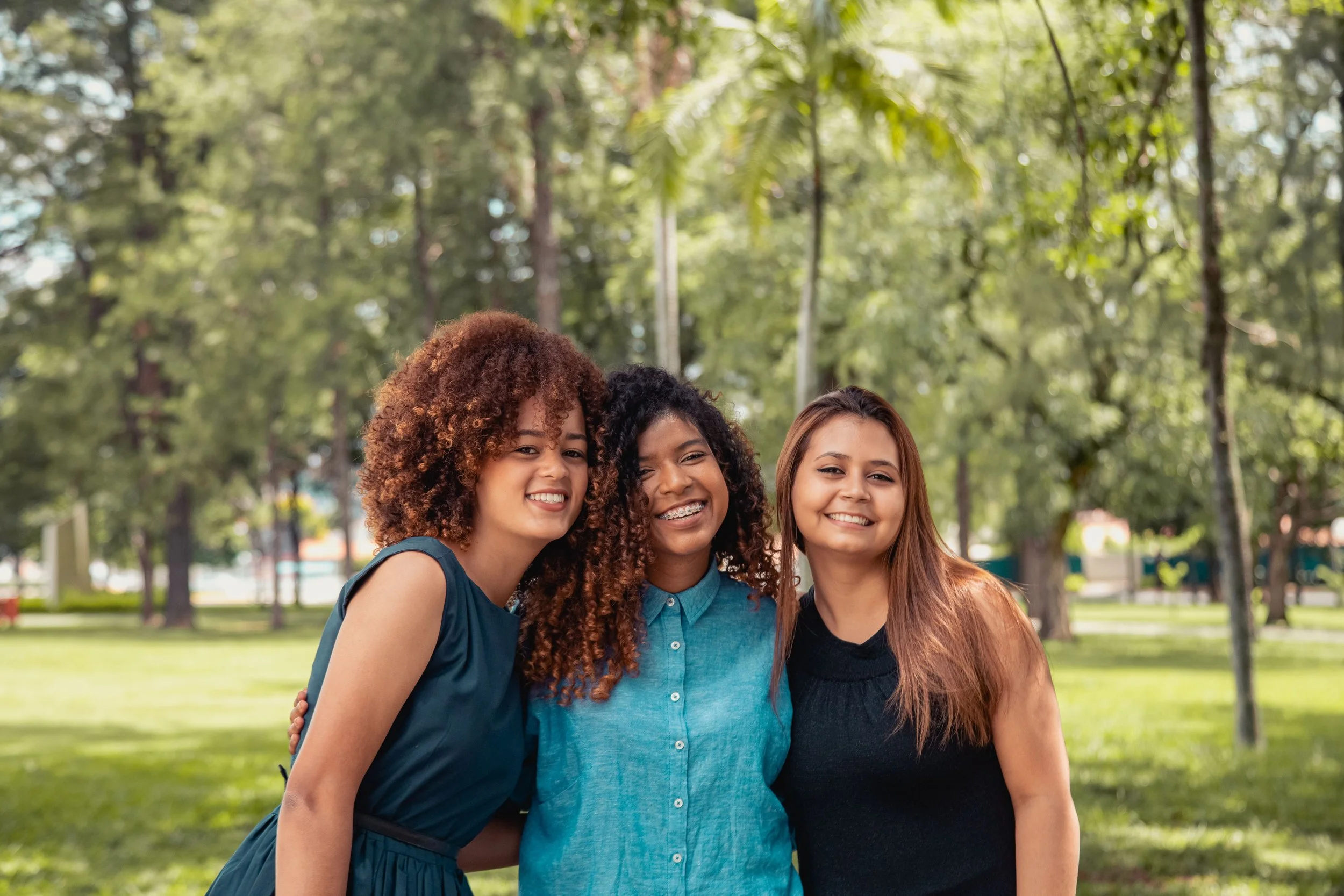 Three young women standing together outdoors in a park, smiling, and embracing each other, with trees and greenery in the background.