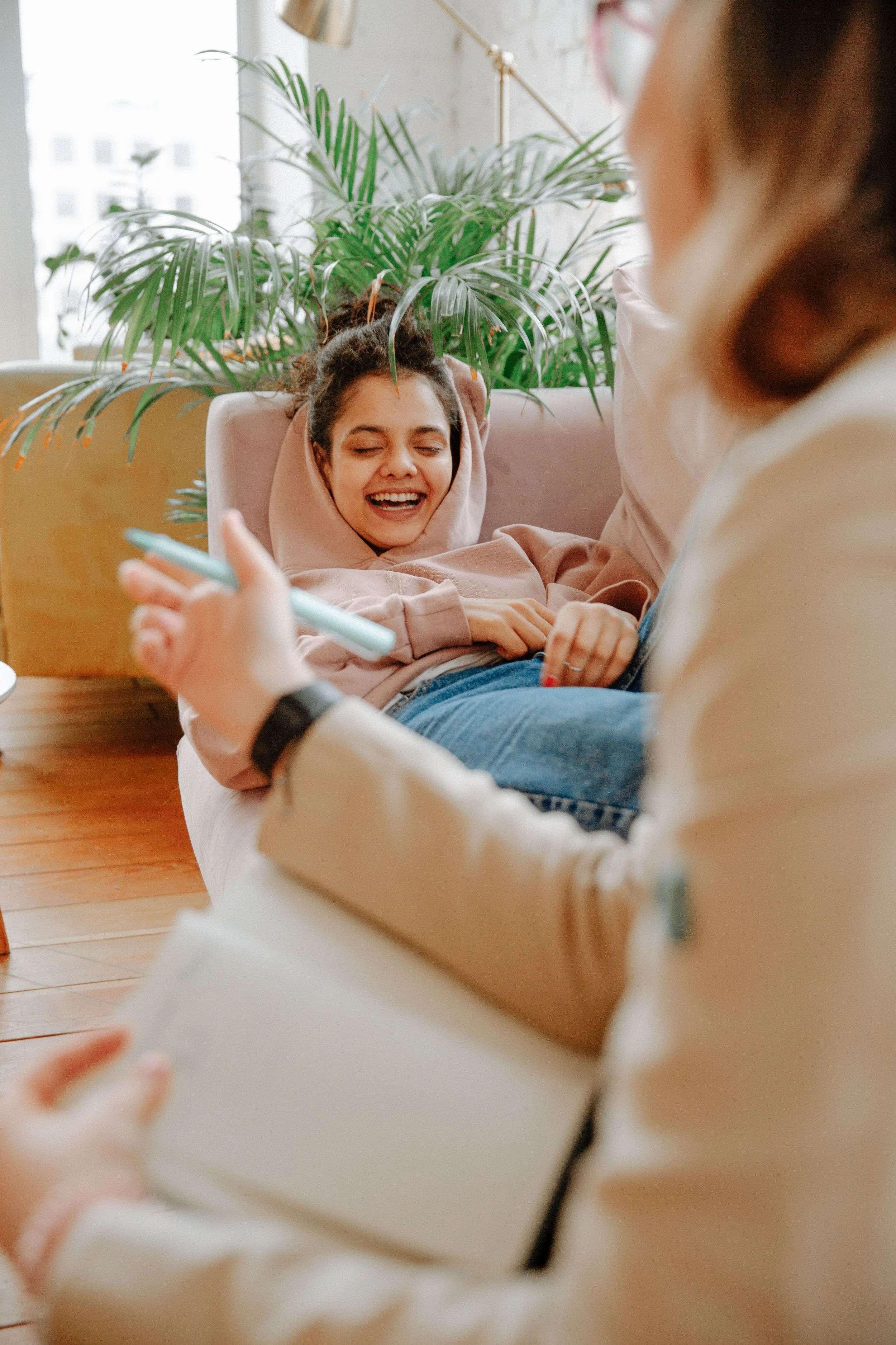 A woman sitting on a light pink couch, smiling and laughing, while another woman in the foreground, partially visible, shows her phone and holds a paper or notebook.