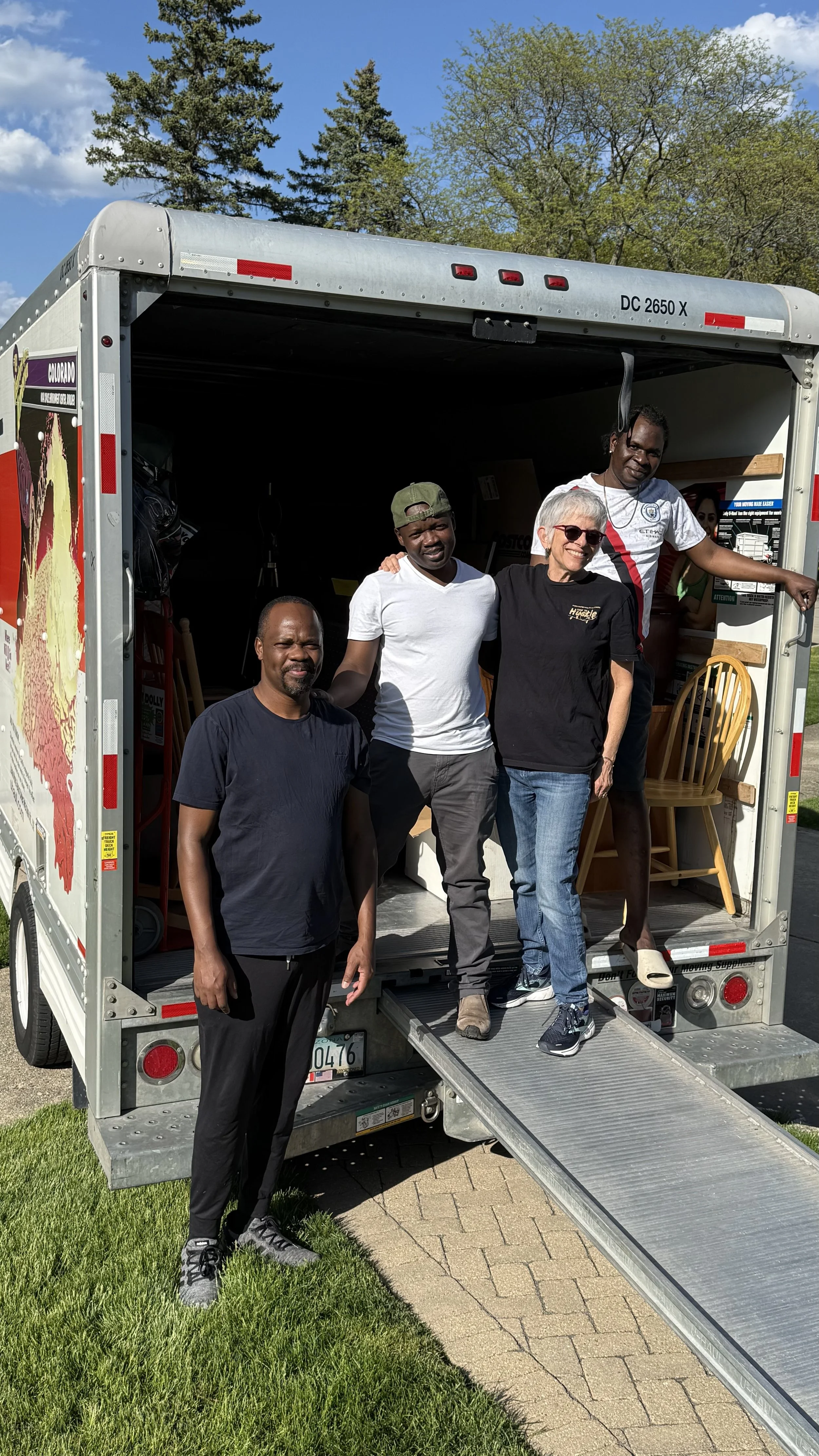 A group of five people standing at the back of a moving truck, with three standing outside and two inside the truck. They are smiling and posing for the photo on a sunny day with a clear blue sky, green trees, and a brick sidewalk.