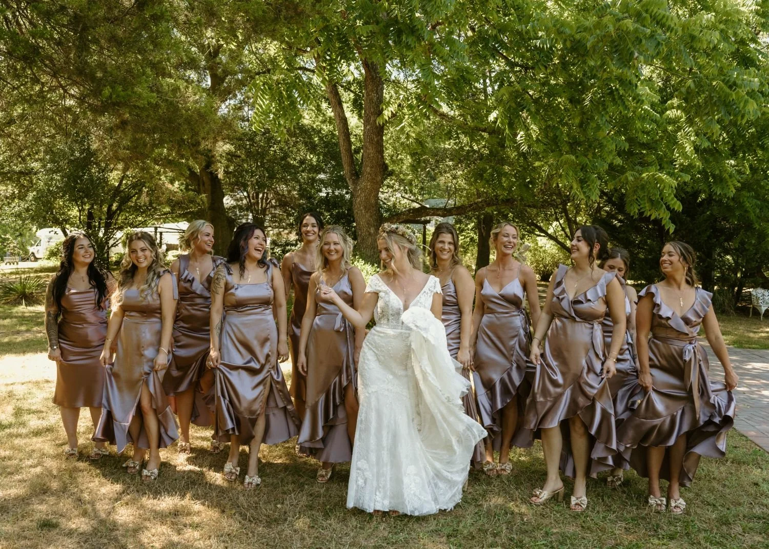 A bride and her bridesmaids dressed in silk gowns walk among the cedar trees before the ceremony at Historic Jasmine, a private wedding venue in New Kent, Virginia.