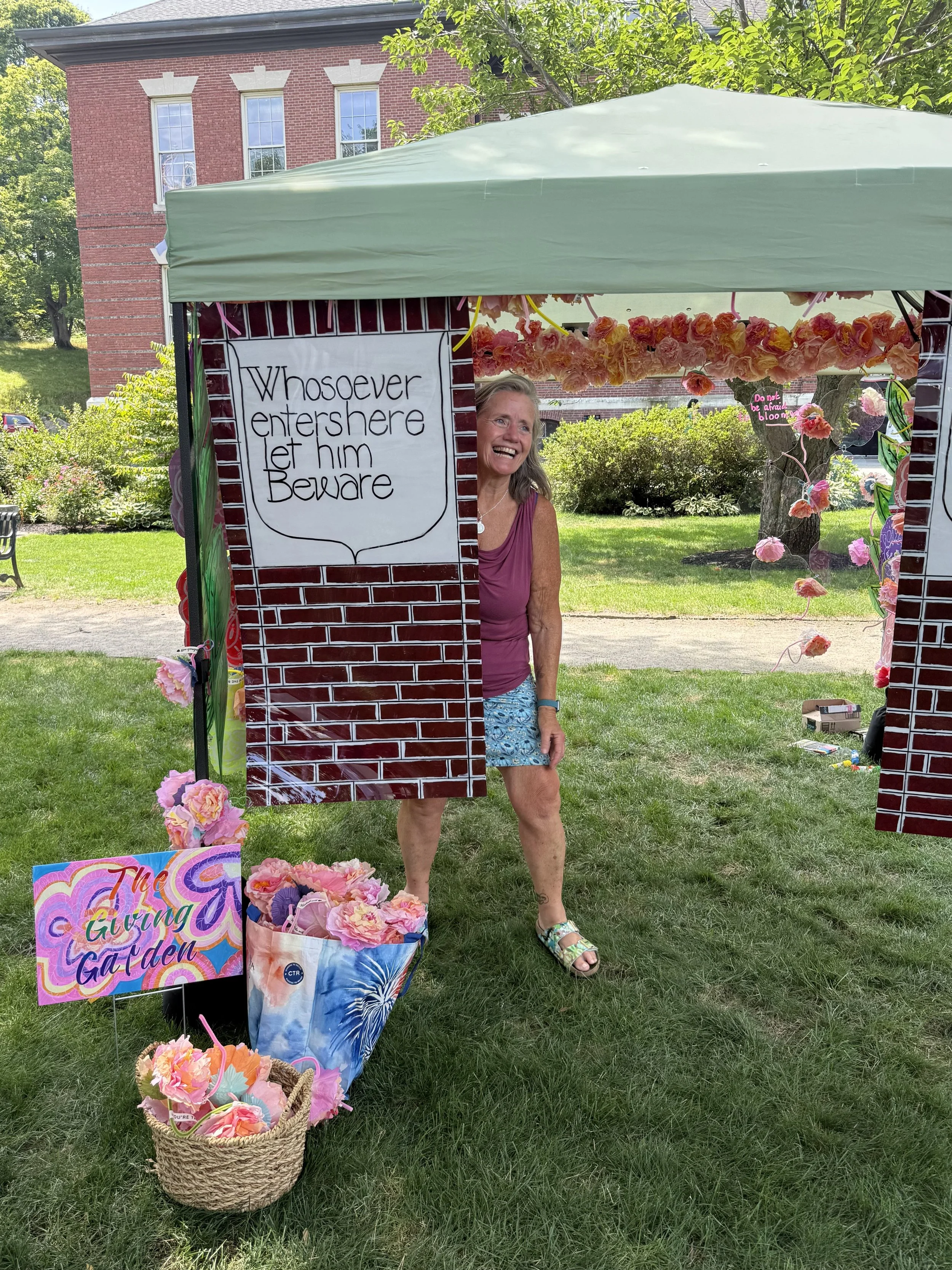 Front view of the installation with a handwritten sign, painted panels, and pink floral elements under a canopy.