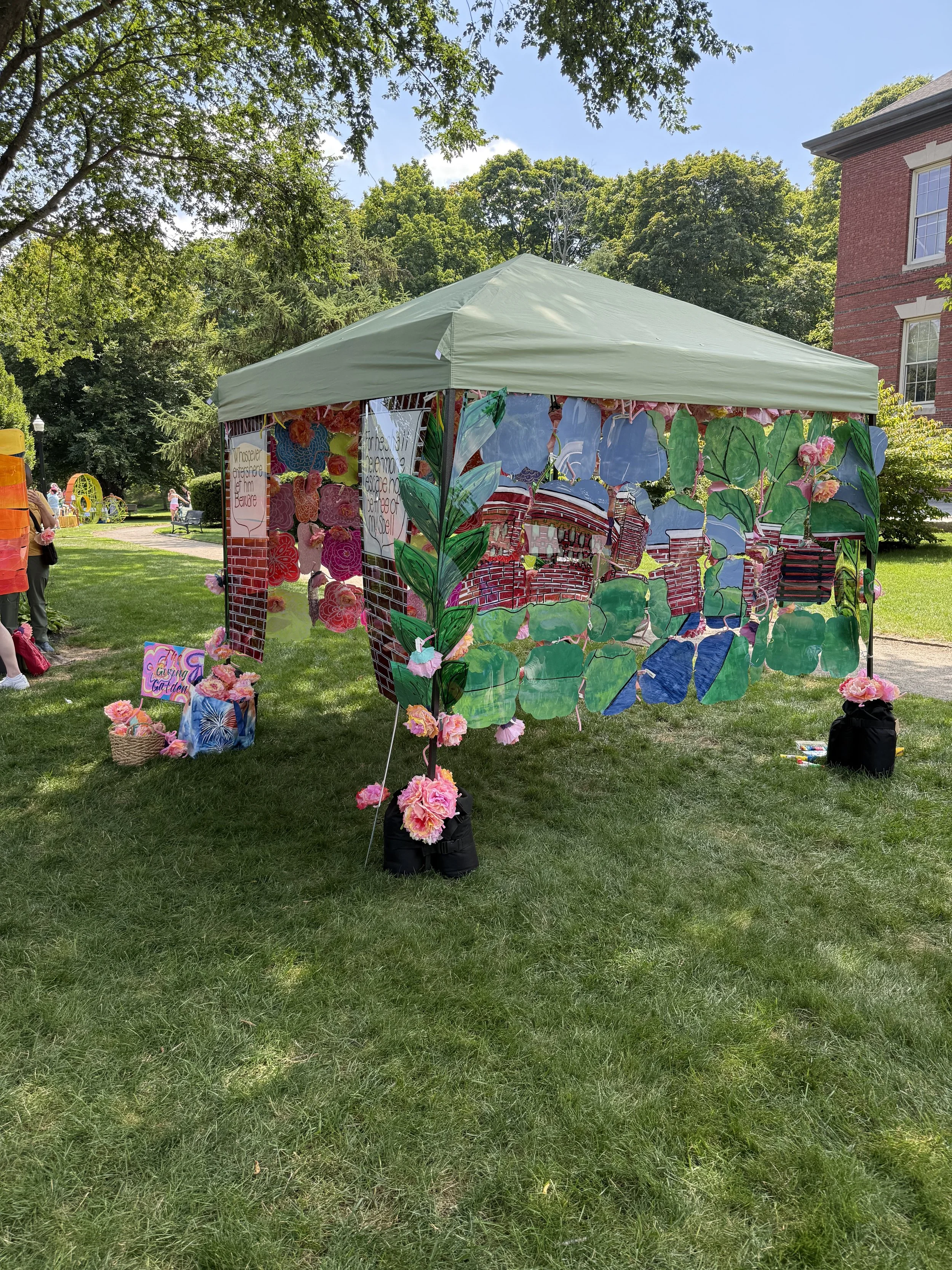 Outdoor installation with a pale green canopy draped in colorful painted translucent panels and pink flower decorations.