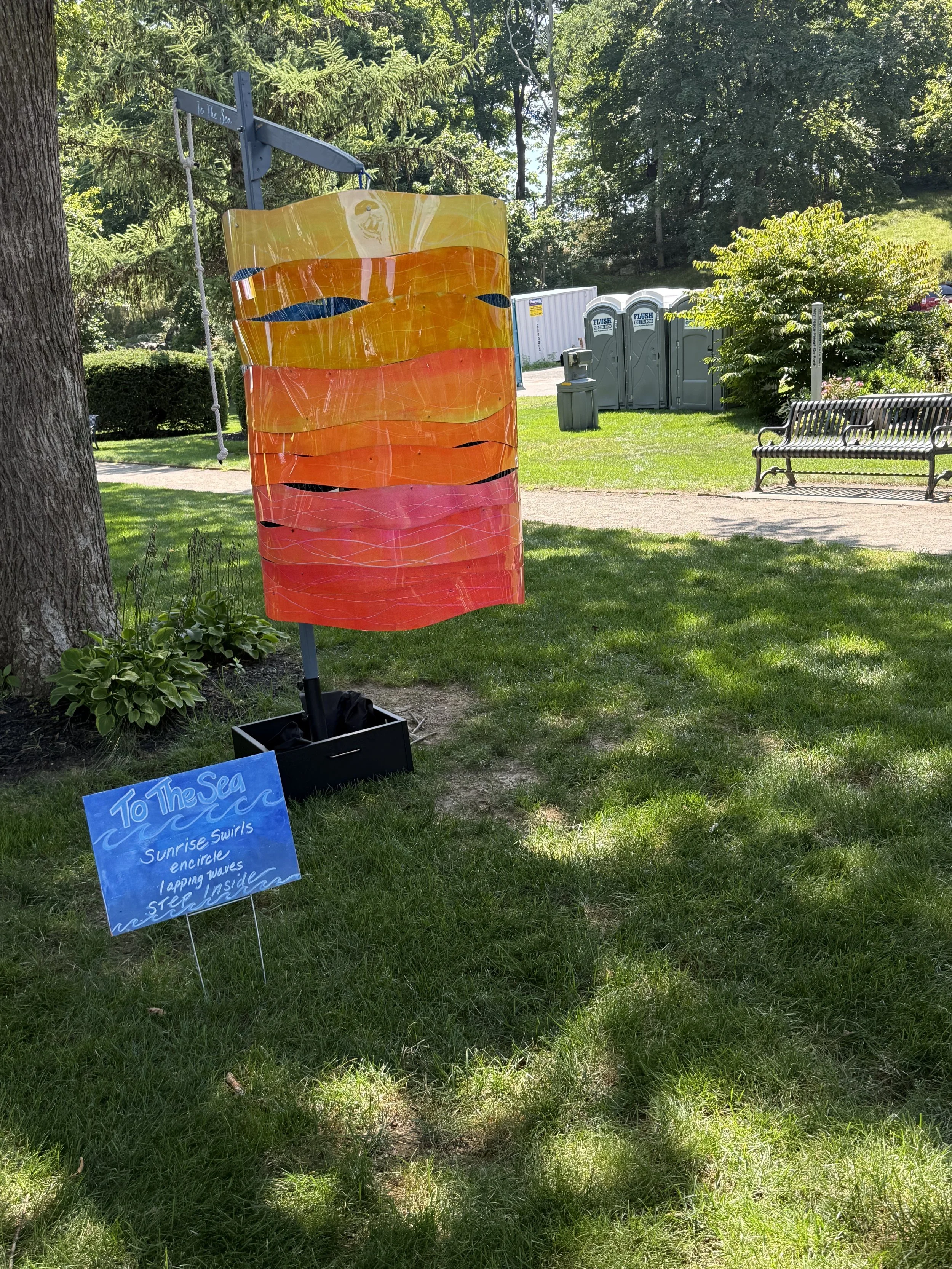 Outdoor view of a freestanding translucent orange-and-pink painted cylinder sculpture beside a tree.