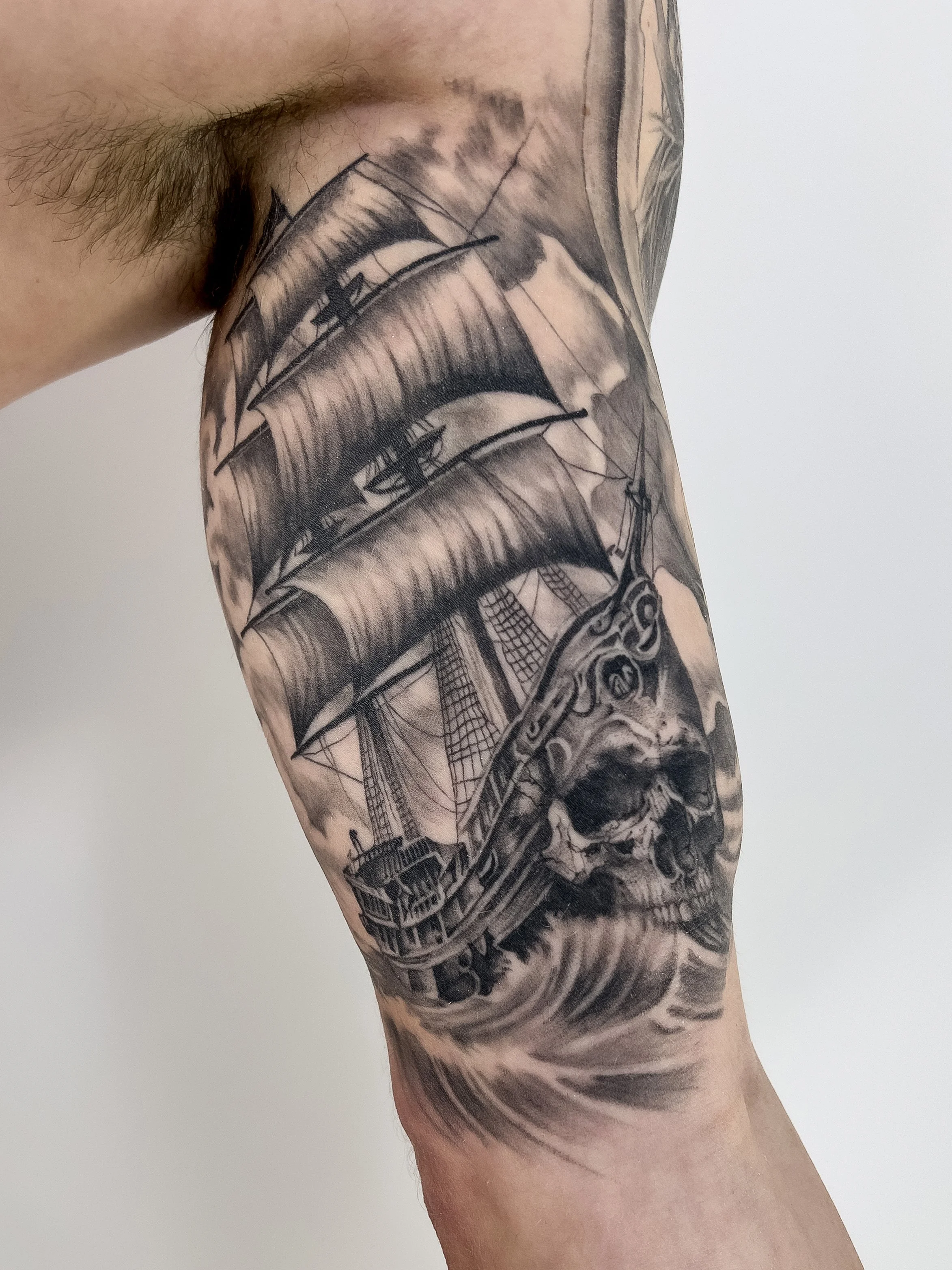 A tattoo of a sailing ship with billowing sails and a detailed skull at the ship's bow on a person's arm.