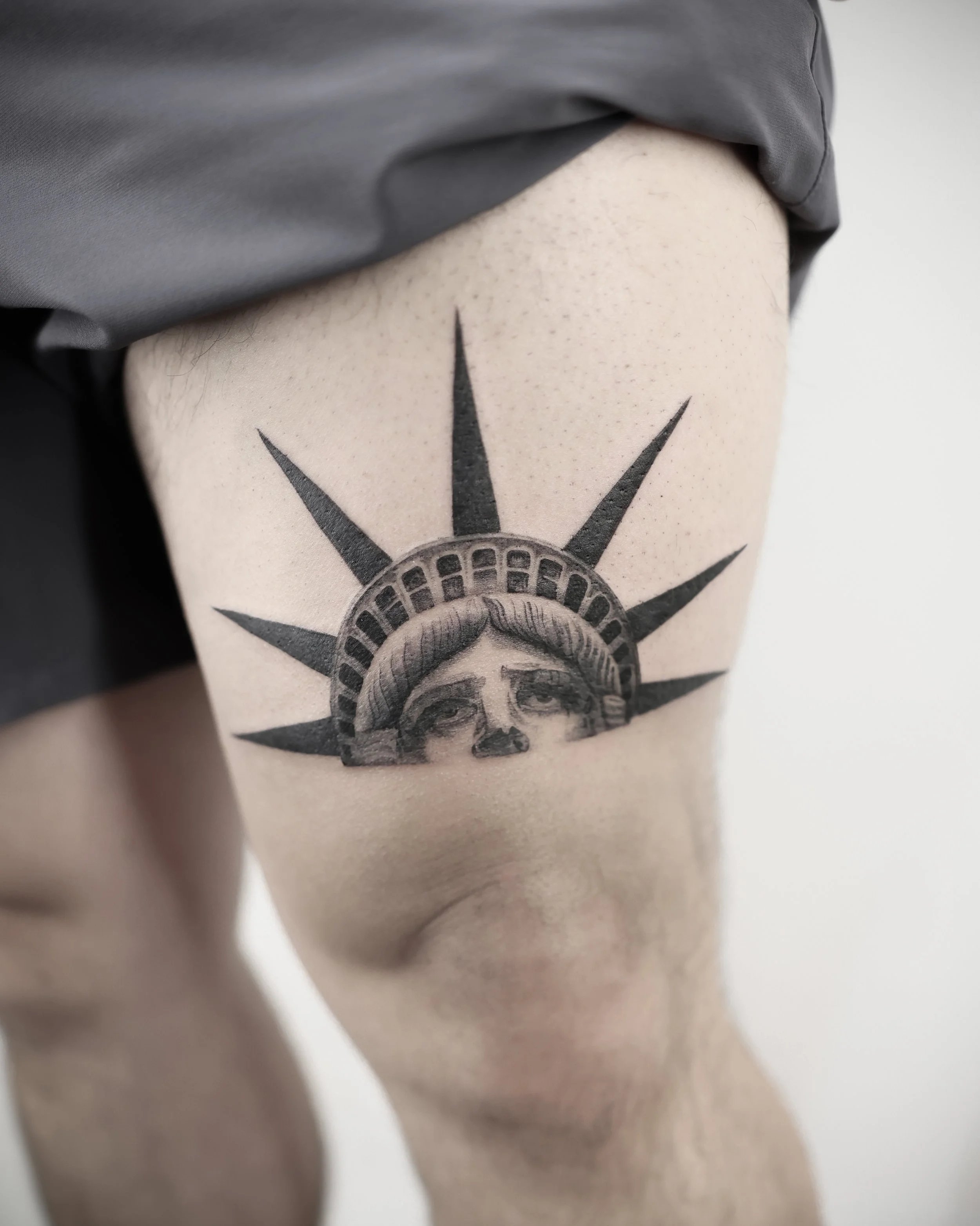Tattoo of the Statue of Liberty's face with the crown's rays extending outward on a person's thigh.