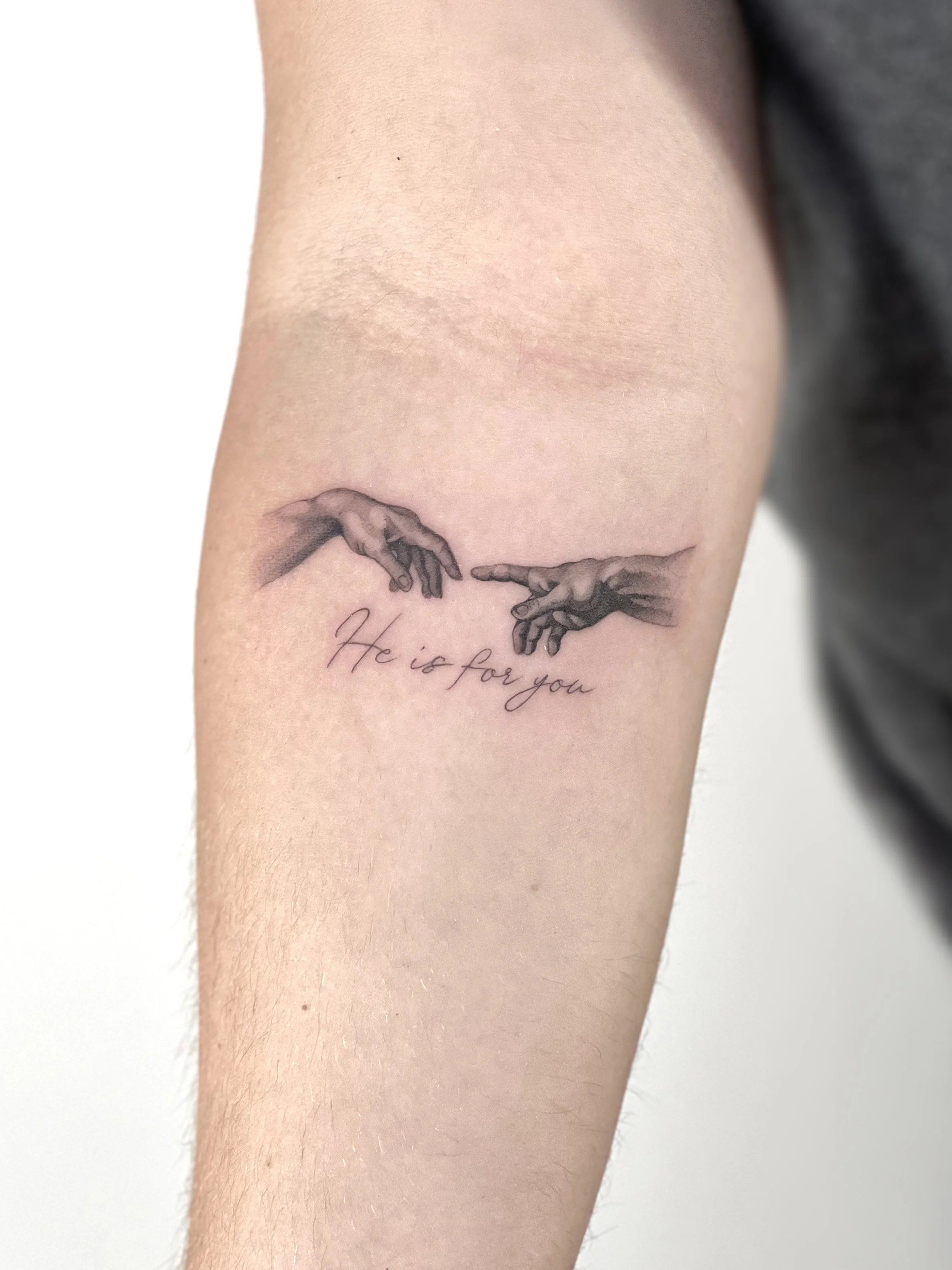A tattoo on the forearm depicting two hands reaching towards each other, with the phrase "He is for you" written below in cursive script.