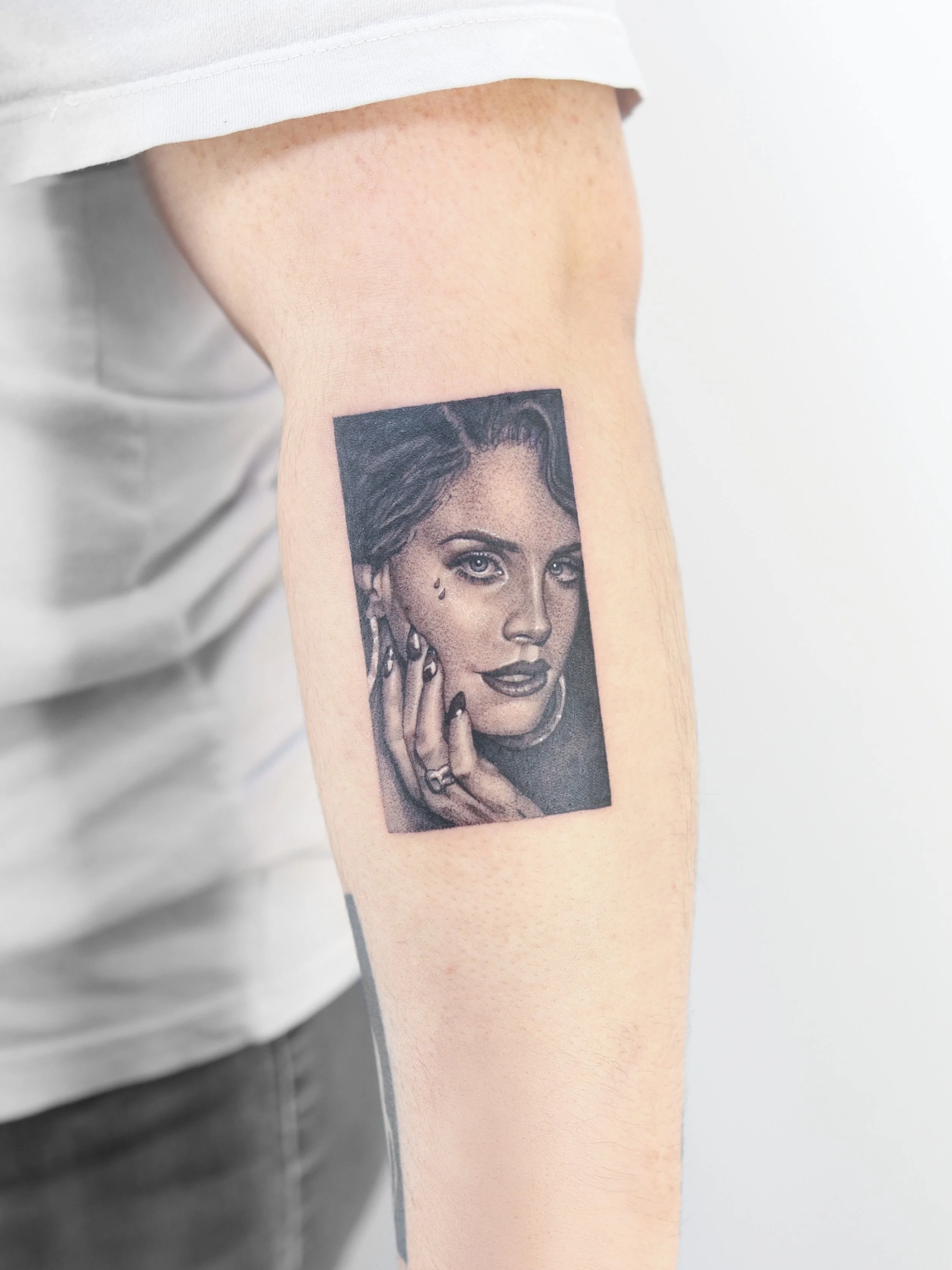 Amazing Black and gray portrait tattoo of a Lana Del Rey with jewelry, pearl earrings, tear near eye, and painted nails, on person's upper arm.