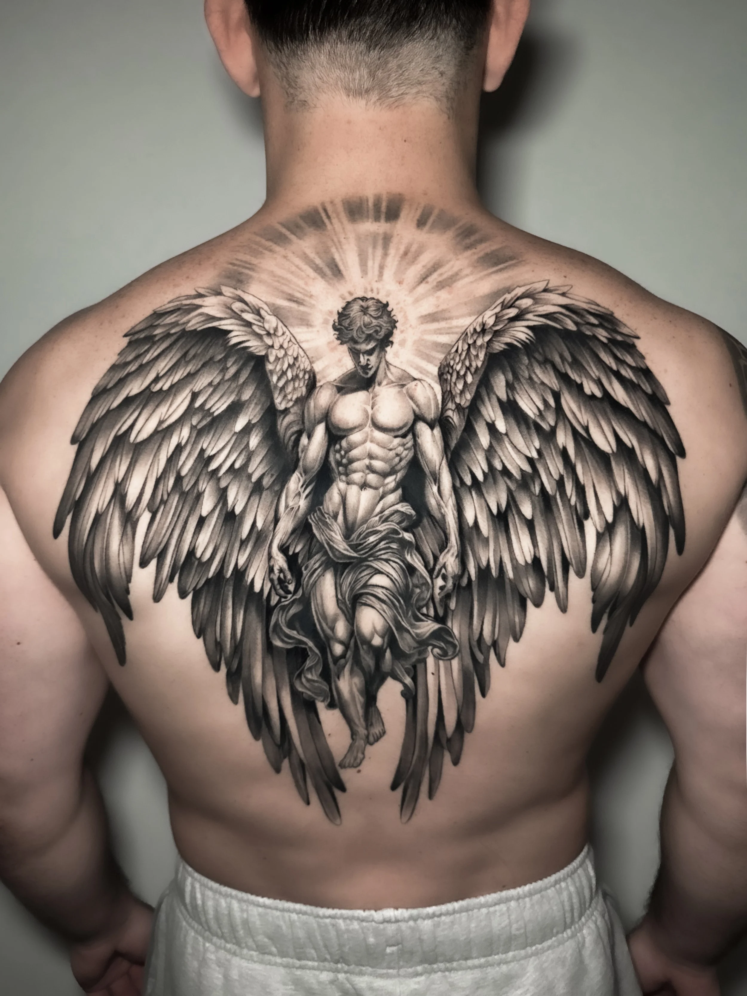 A detailed black and gray tattoo of an angel with large wings on a person's back, with a halo radiating light behind the angel's head.